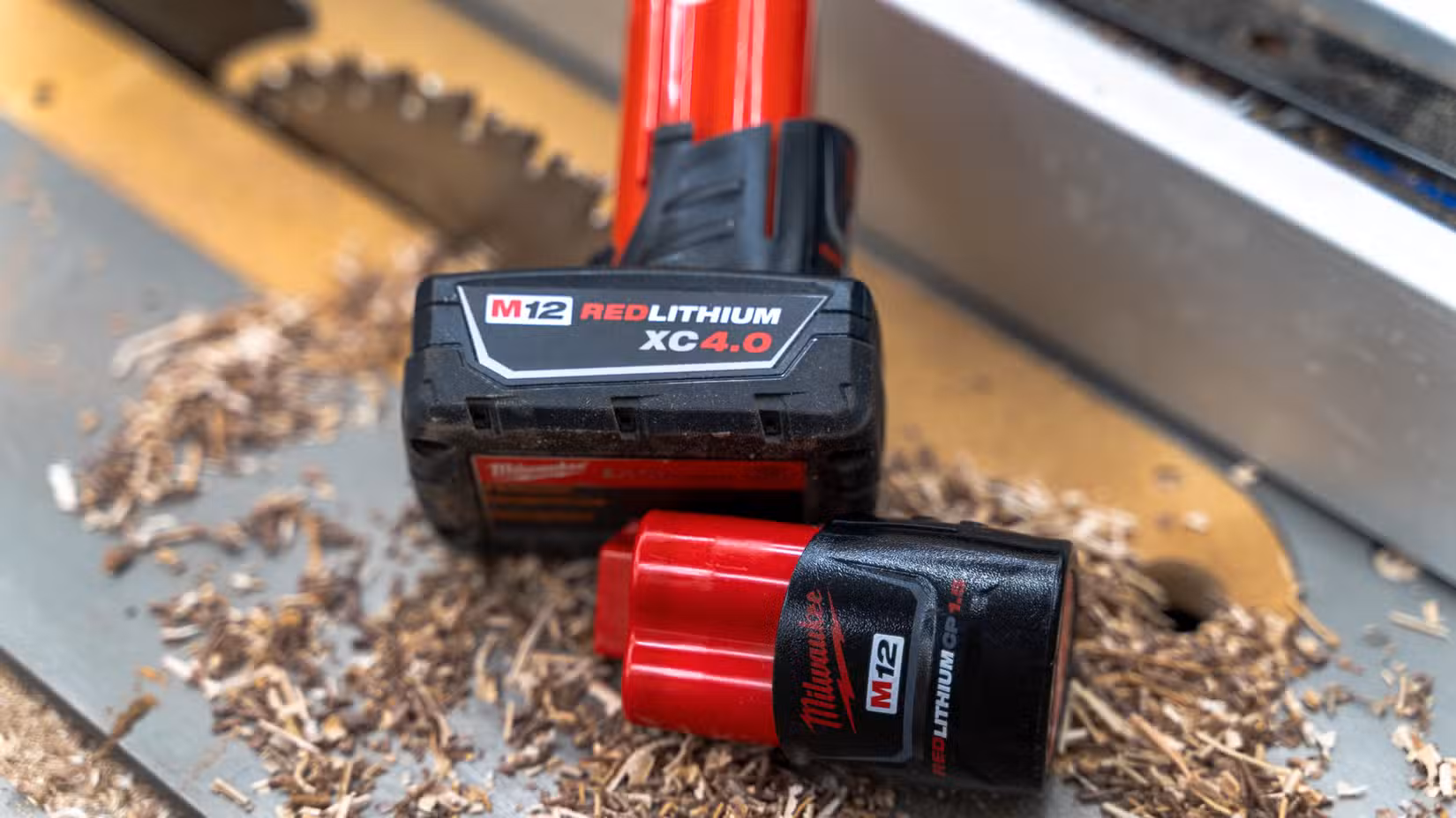 Two Milwaukee M12 batteries sitting on a table saw with sawdust and a blade in the background.