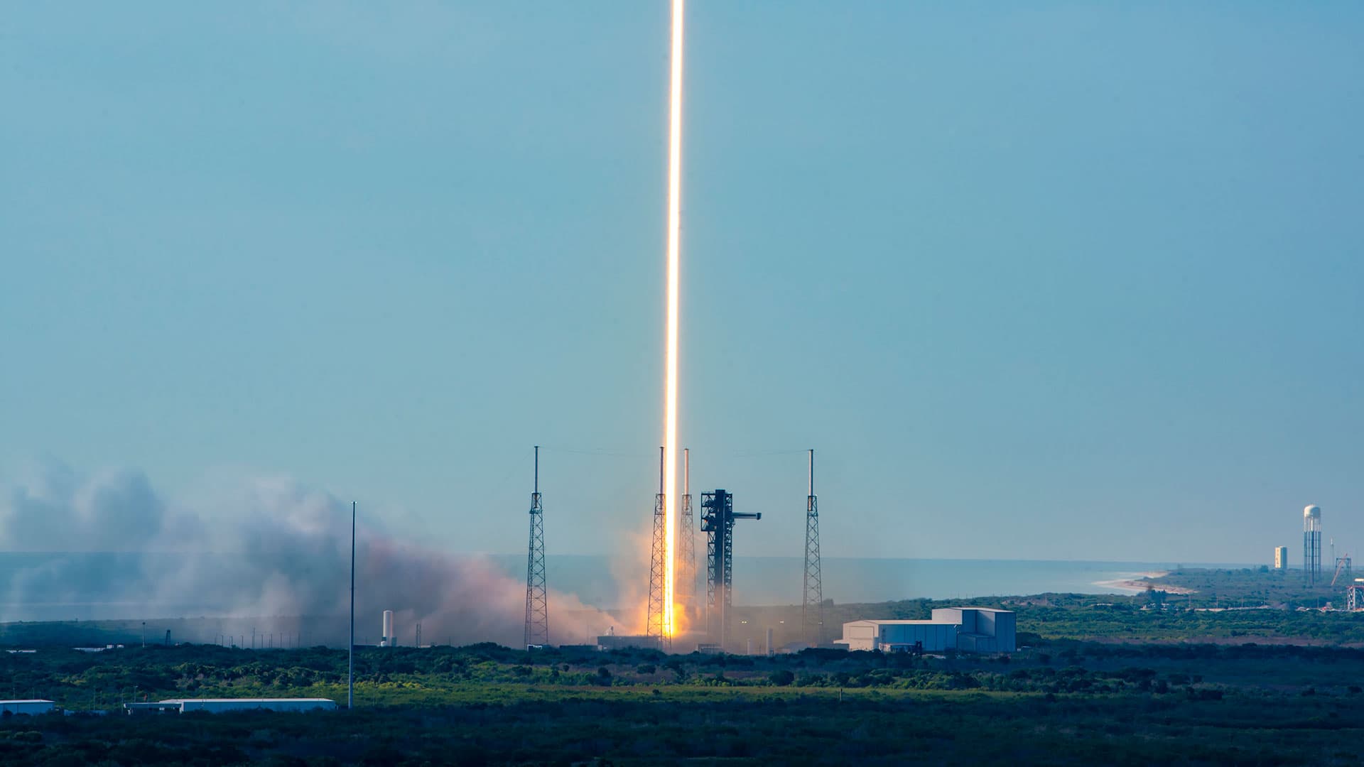 A time lapse photo of a daytime rocket launch shows the rocket's fiery plume as a single orange and white beam heading skyward off the launch pad into the blue sky.