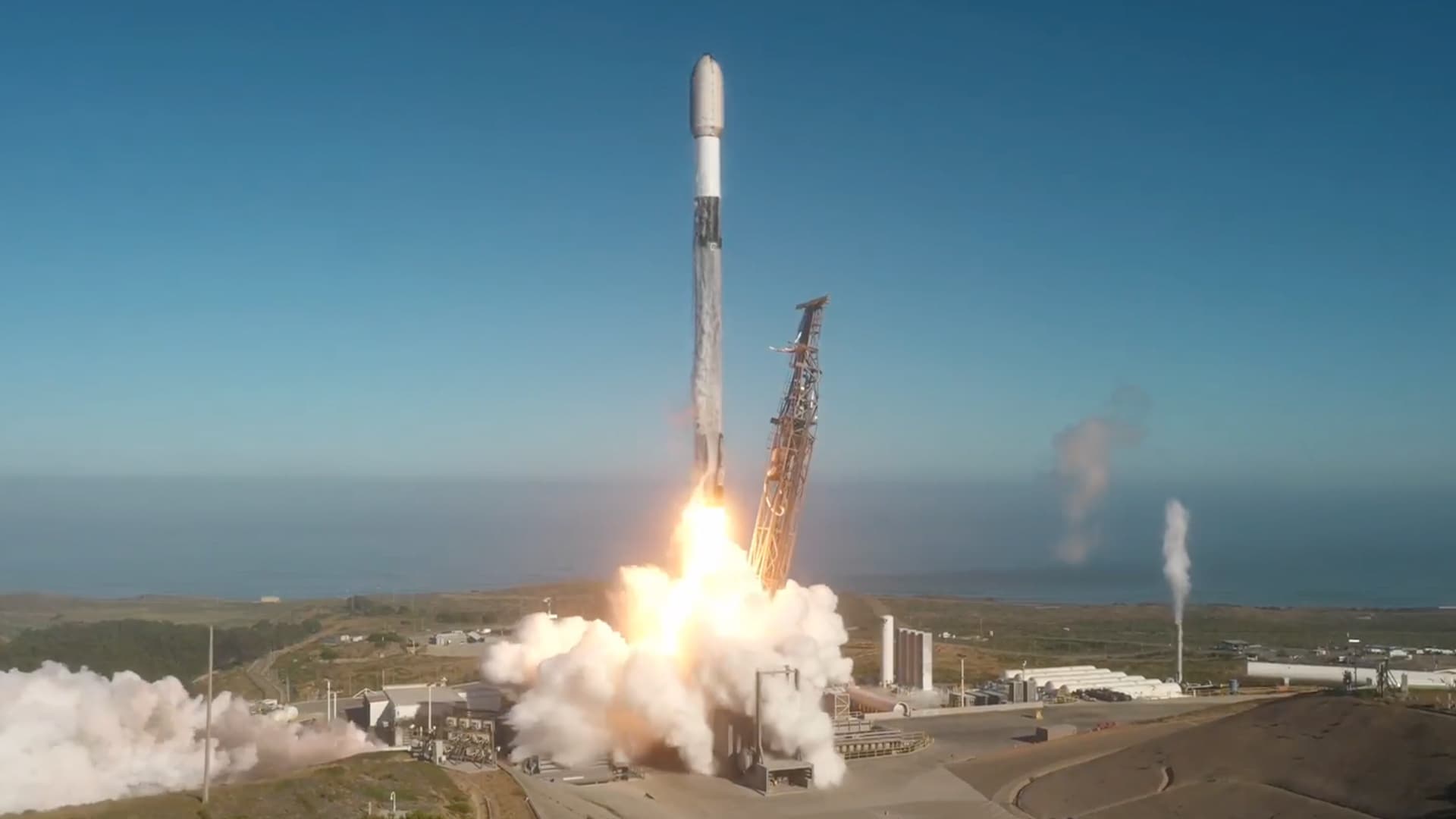A white, black and gray rocket lifts off into a blue sky from an oceanfront launch pad.