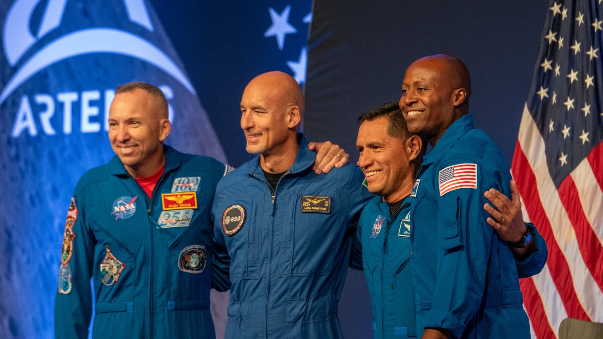 four people in blue flight suits smile and embrace one another on stage