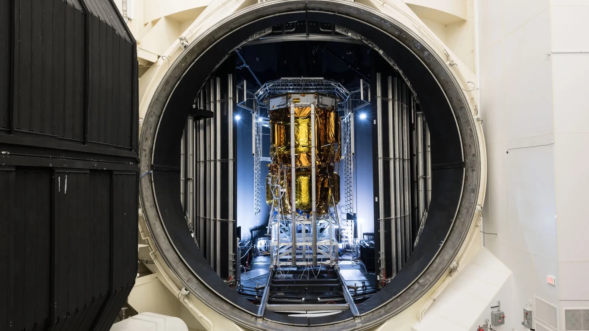 a gold-foiled spacecraft stands in a chamber with an open hatch