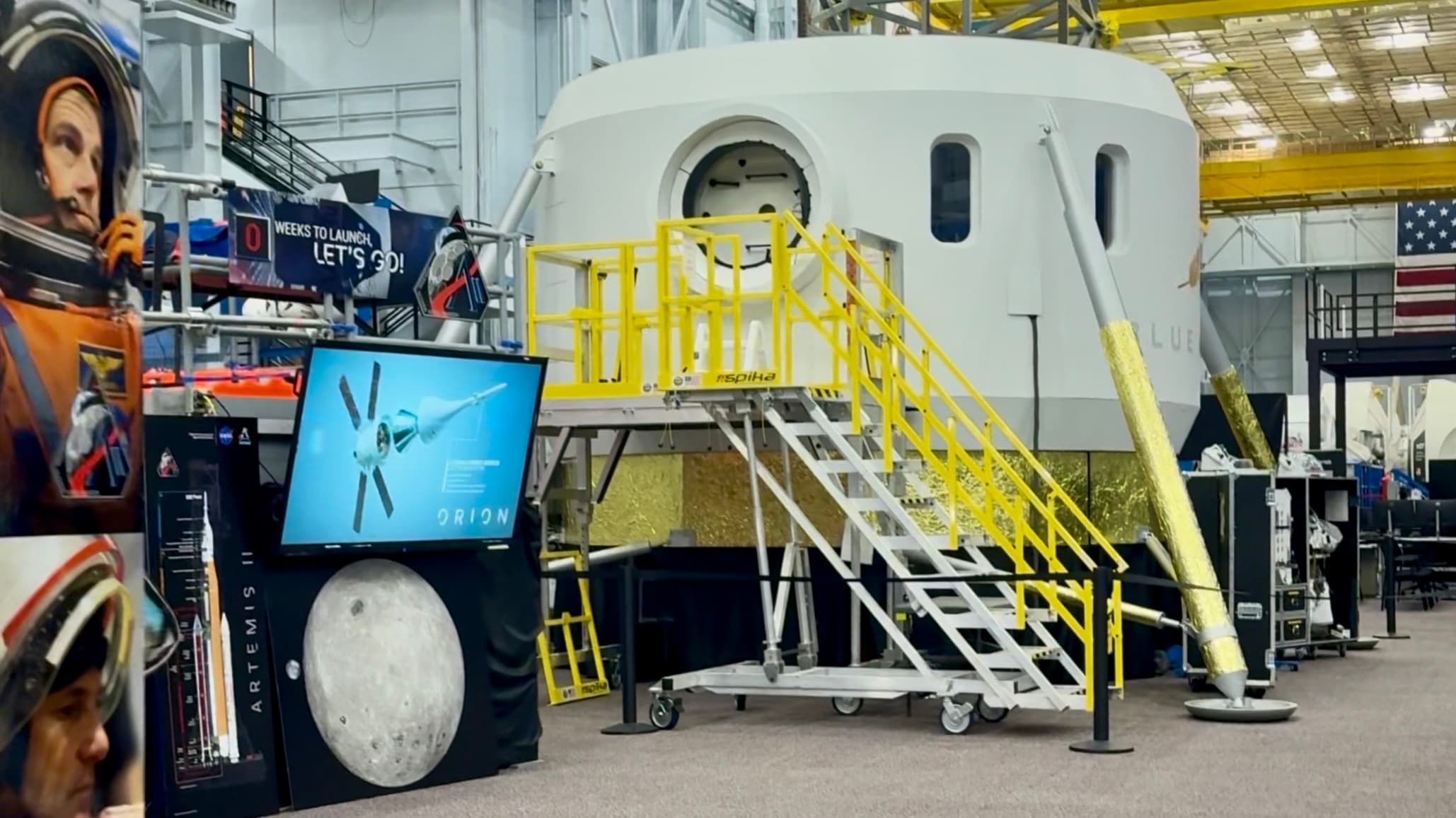 a cylindrical white capsule stands on golden-foil-wrapped legs with yellow scaffolding steps against a rounded hatch inside a big spacecraft mockup training facility.