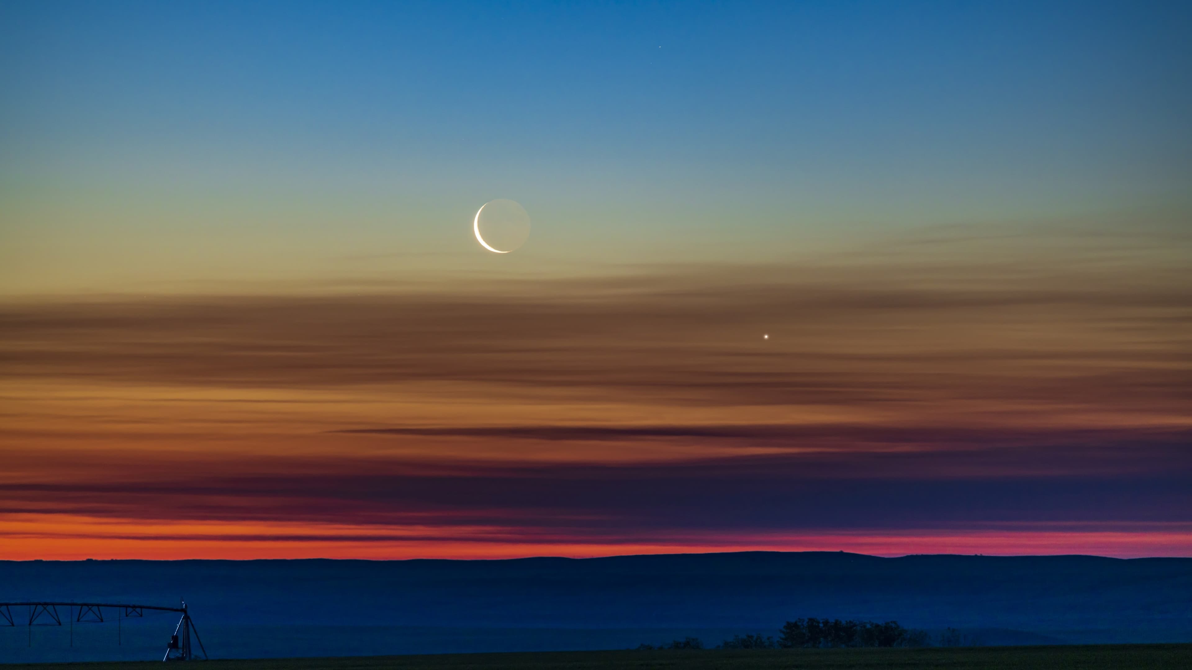 the moon shines next to bright venus at sunset. the sky is painted hues of blue, yellow and orange.