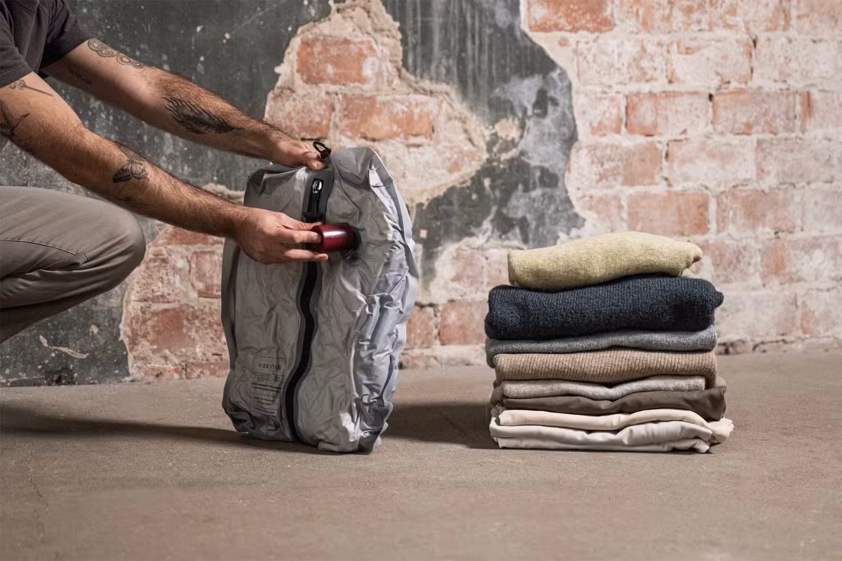 A man's hand compressing an Ekster vacuum packed travel bag with a pile of sweaters beside it.