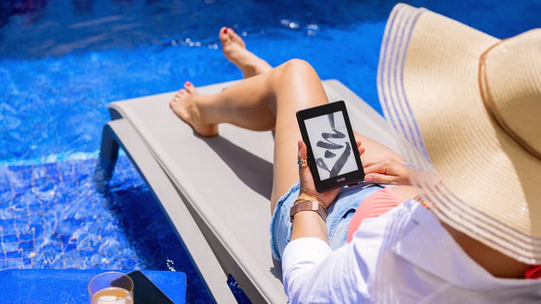 A woman in a sun hat lounges on a pool in the shallow end of a pool with a Kindle on her lap.