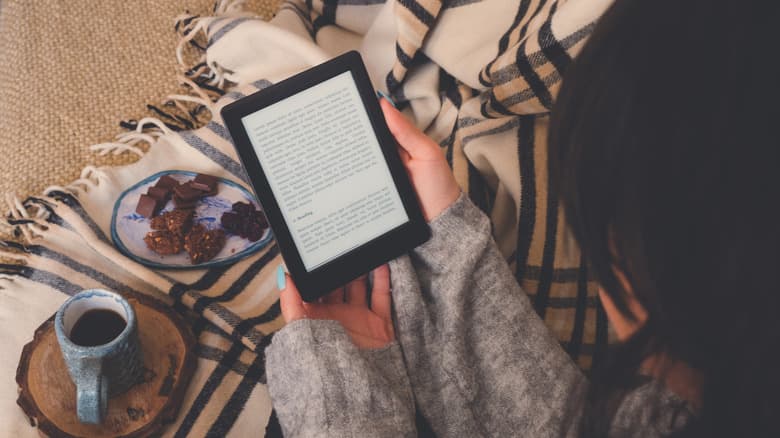 A woman sitting on a blanket reads from her Kindle, sampling a cup of coffee and a plate of treats.
