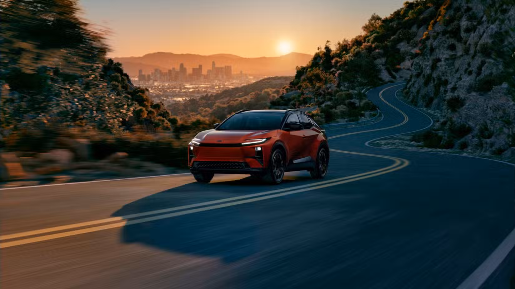 Dynamic front 3/4 shot of a red 2026 Toyota C-HR driving up a winding road with a city in the background at sunset.