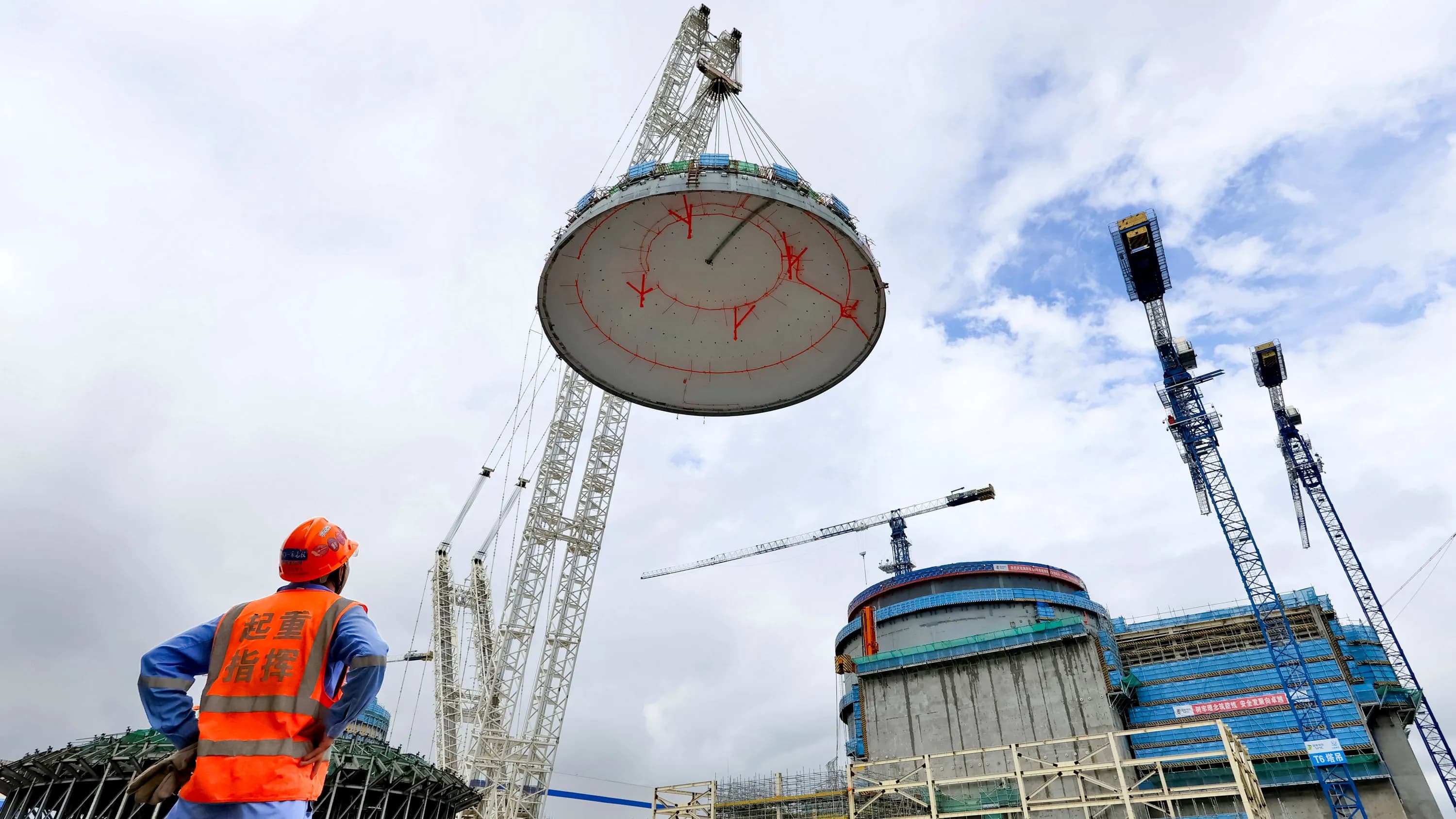 A worker in the foreground looks up at the dome as it is hoisted by crane to the containment vessel of a nuclear power plant