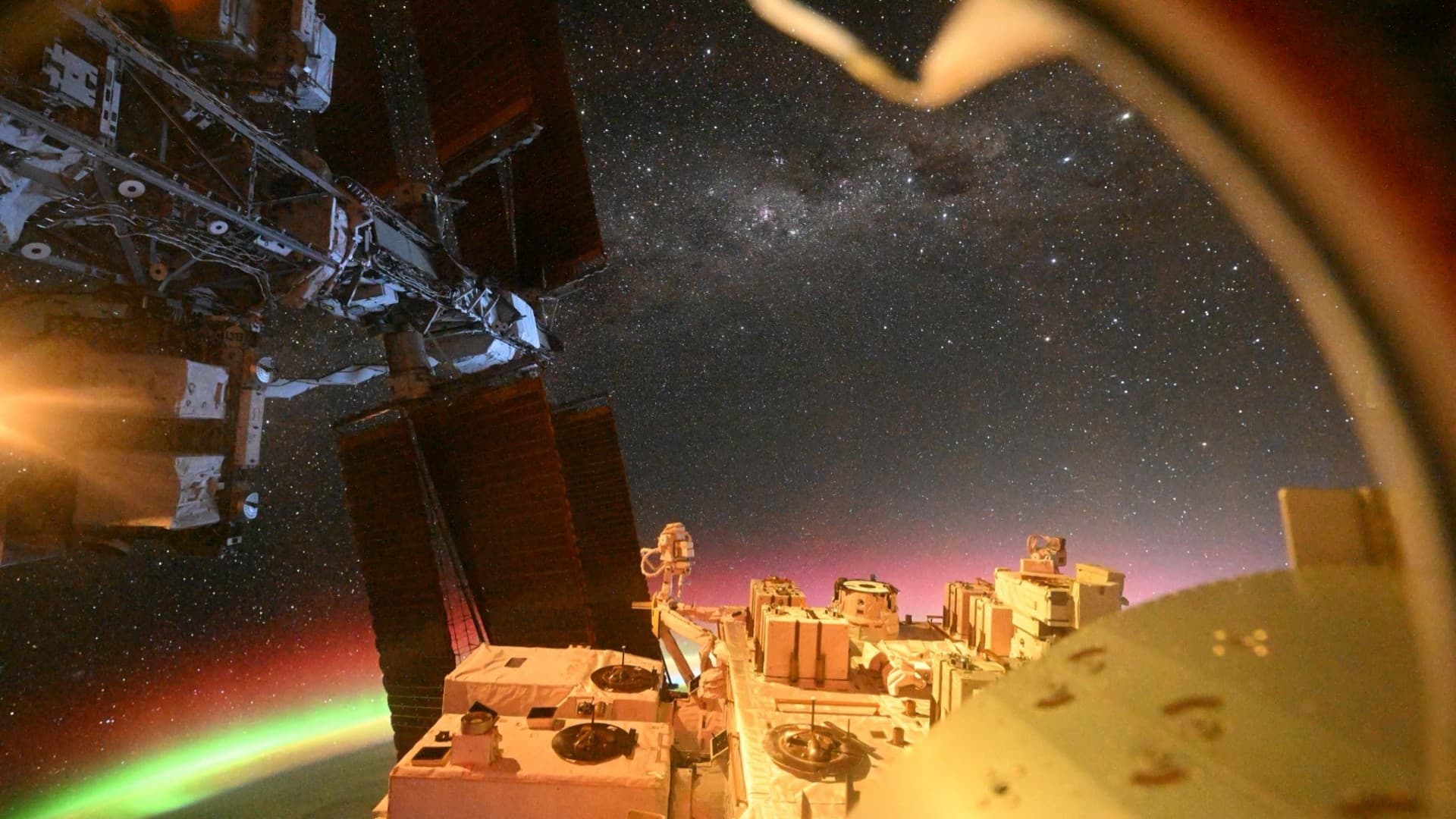 A view from the ISS that shows the ISS solar panels, auroras over Earth, and outer space with a vast array of stars.