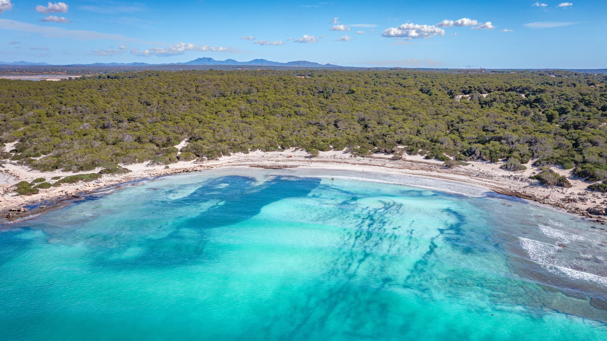 aerial view of a white sand beach and azure blue waters and sky above. Behind the sanding beach is vast swathes of greenery with some mountains in the far distance.