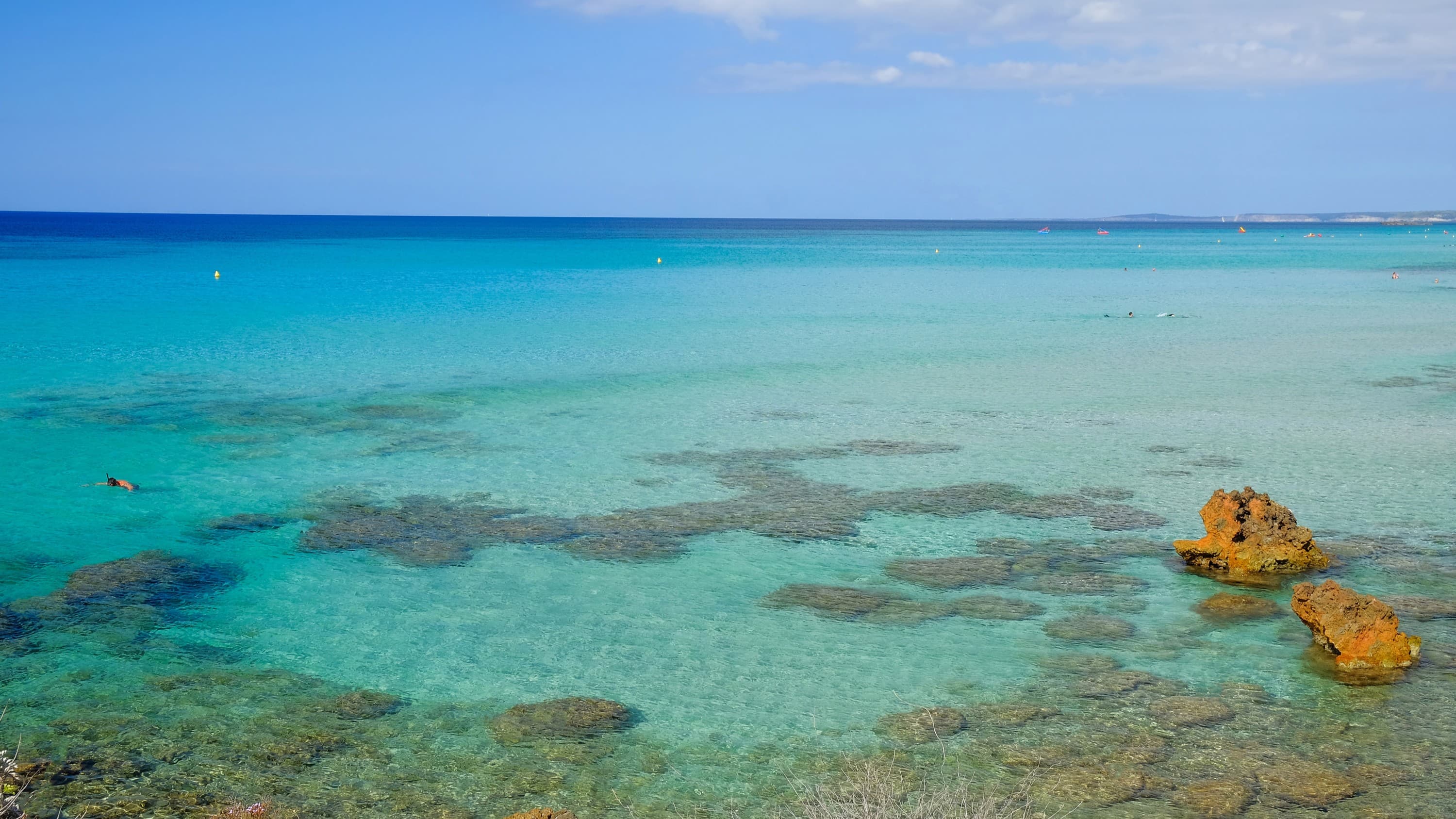 stunning blue green ocean with patches of rocky outcrops, above is a light blue sky with a handful of white fluffy clouds.