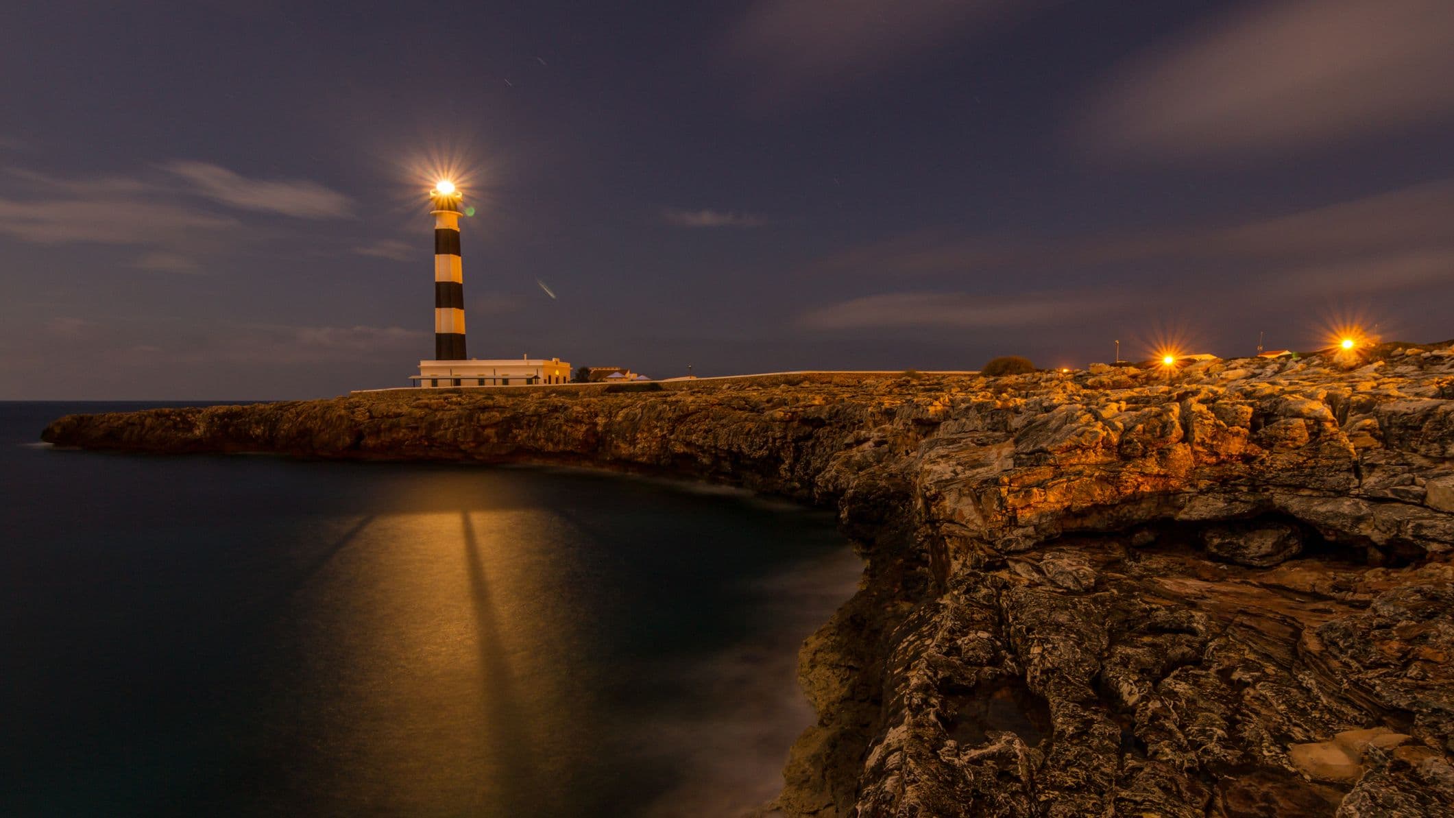 night scene showing a stripey light house on the edge of a rocky outcrop