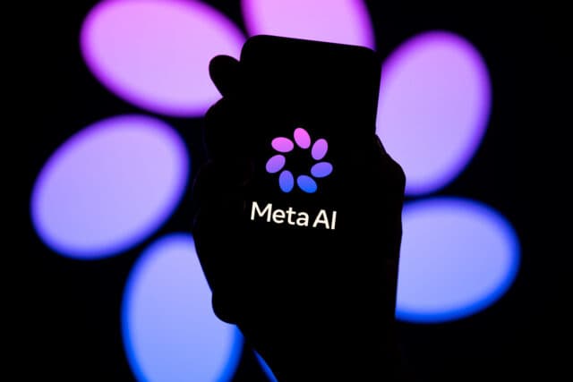 The words "Meta AI" in white are seen with a purple petal pattern logo for Meta AI on a dark smartphone screen. A dark hand holds the phone. The background shows a bigger version of the Meta AI logo.
