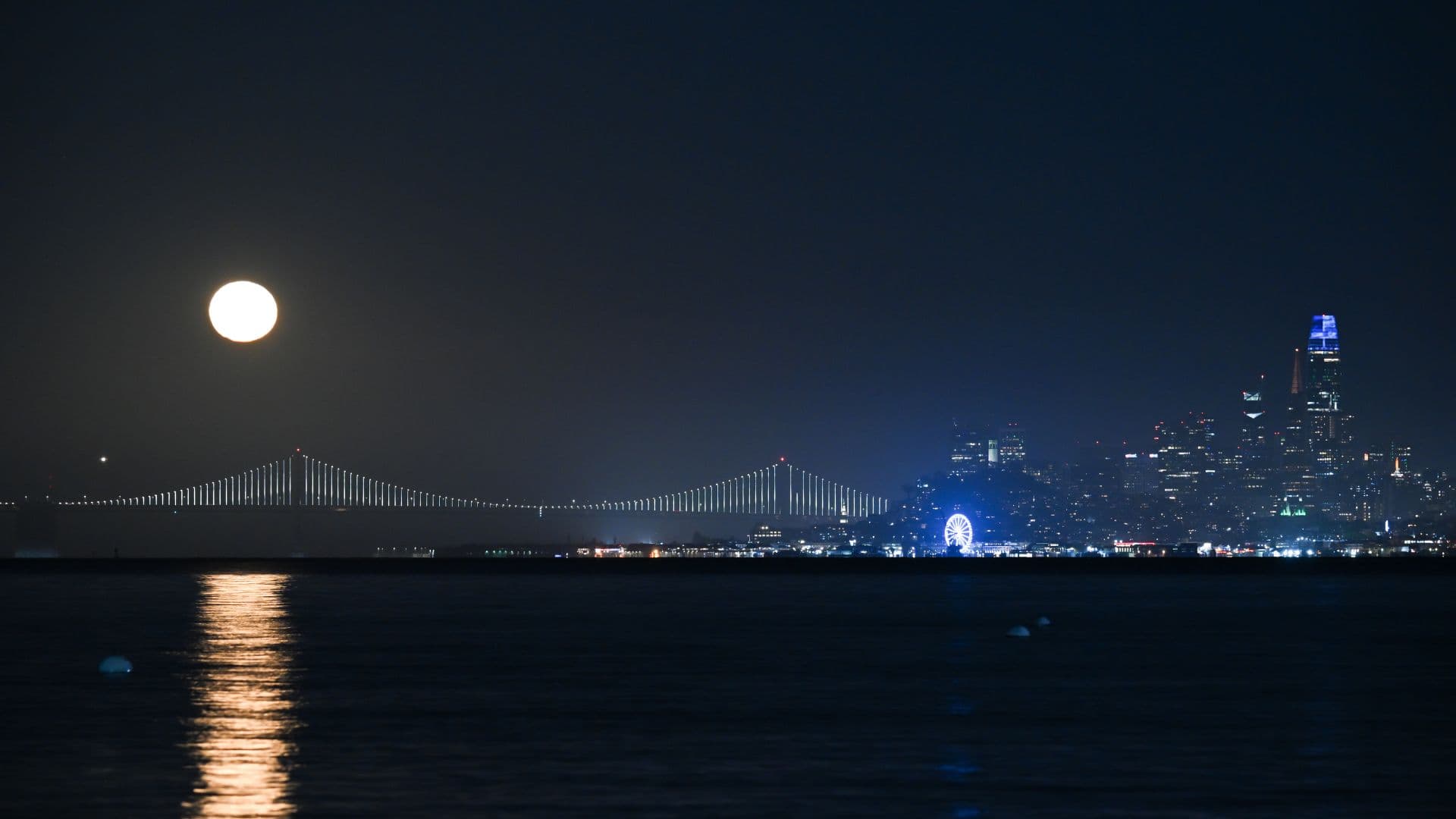 A full moon rises in a night sky above an illuminated suspension bridge, as a city skyline towers to the right. A body of water reflects the moonlight in the foreground.
