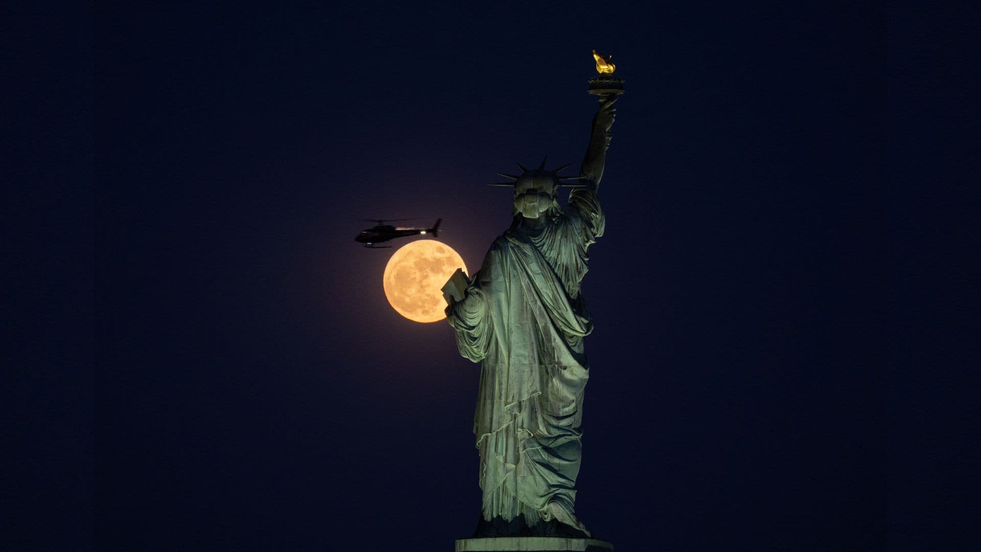 A yellow full moon glows behind the Statue of Liberty in New York at night, as a helicopter passes above.