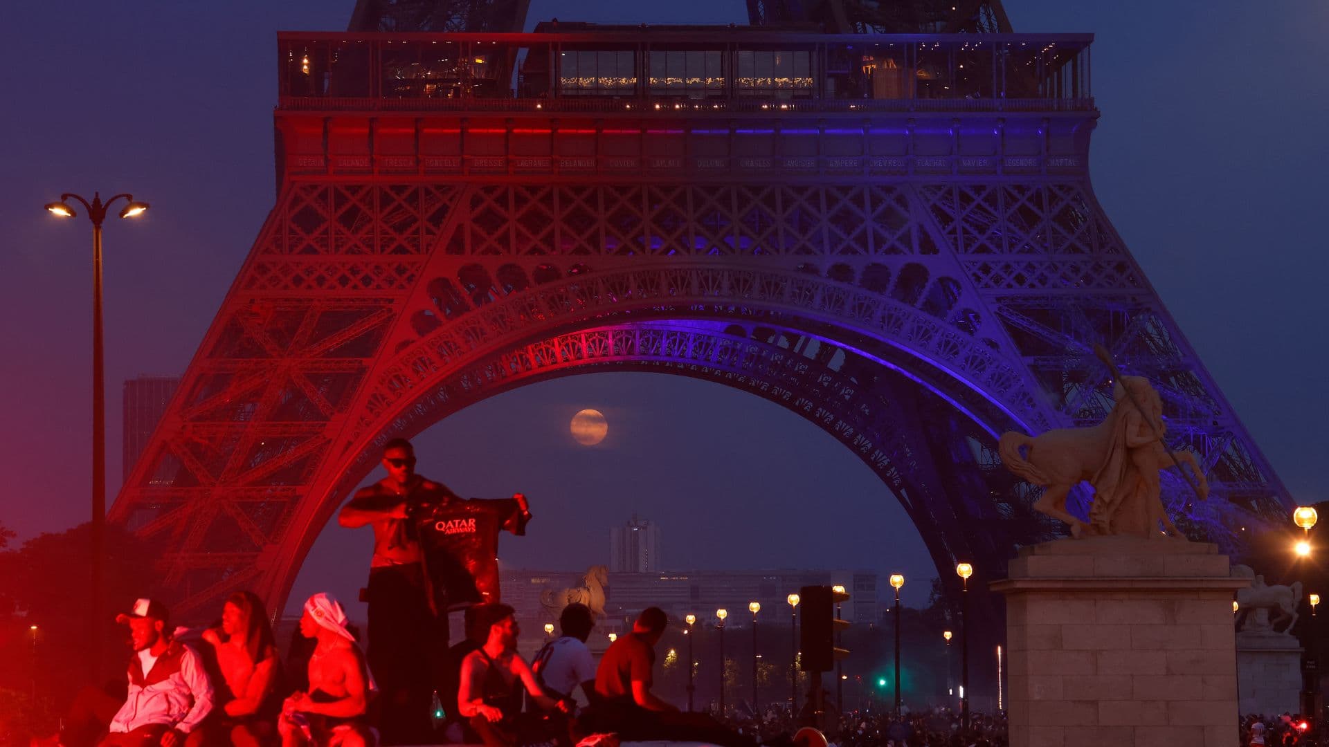 Fans celebrate as the full moon rises between the steel legs of the Eiffel Tower, which is lit in red and blue.