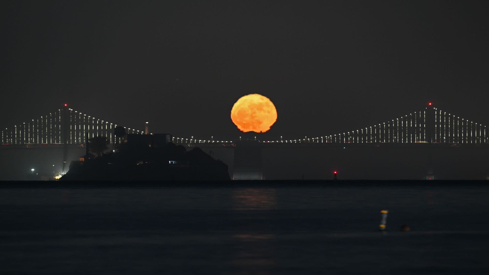 A yellow full moon shines in a dark sky above an illuminated suspension bridge above a dark body of water.