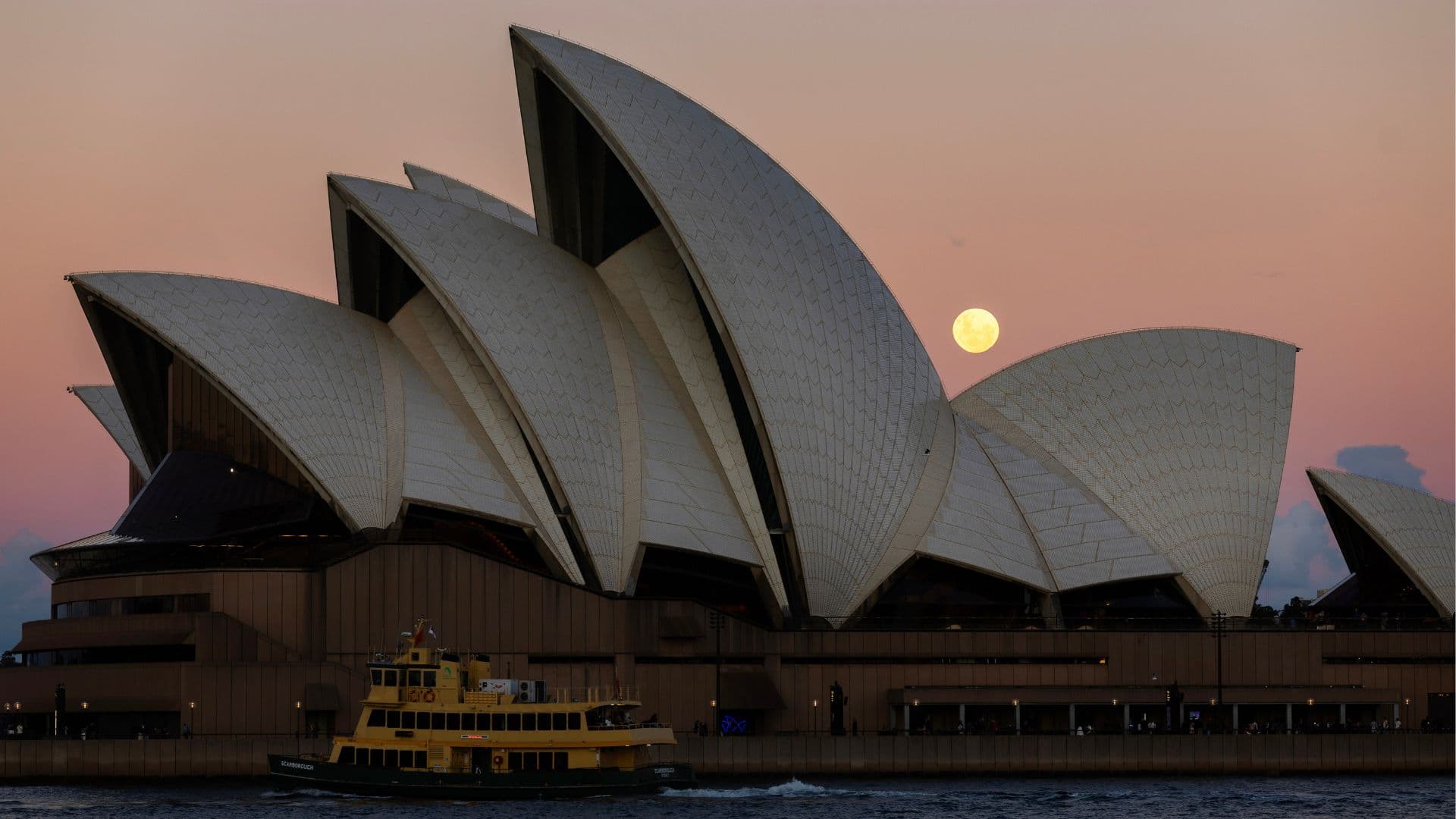 A yellow moon rises above the Sydney Opera House in a peach sky.