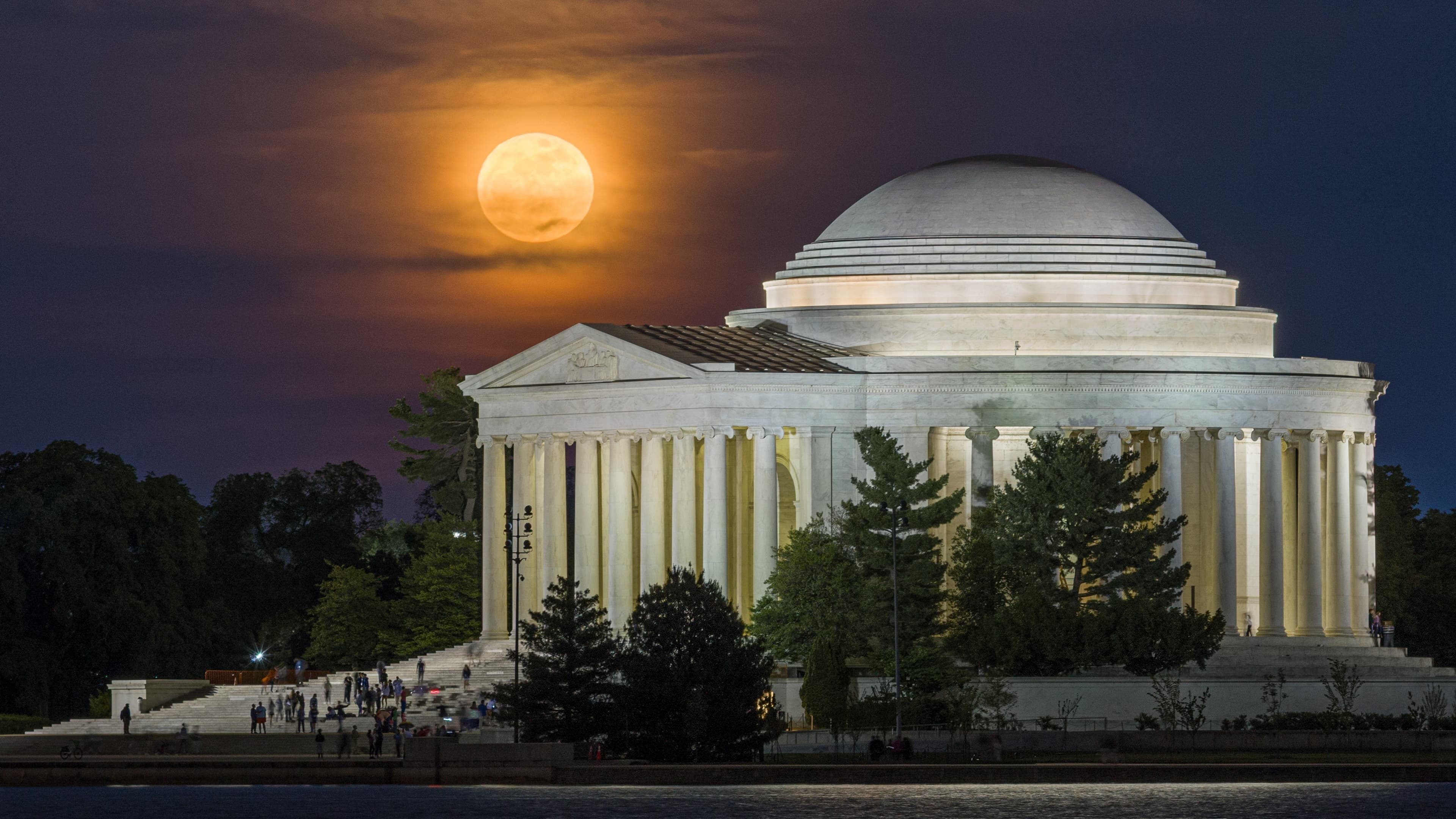 An orange full moon illuminates thin clouds above the domed exterior of the Jefferson Memorial in Washington D.C. at night. Trees are visible in the foreground bordering a lake.