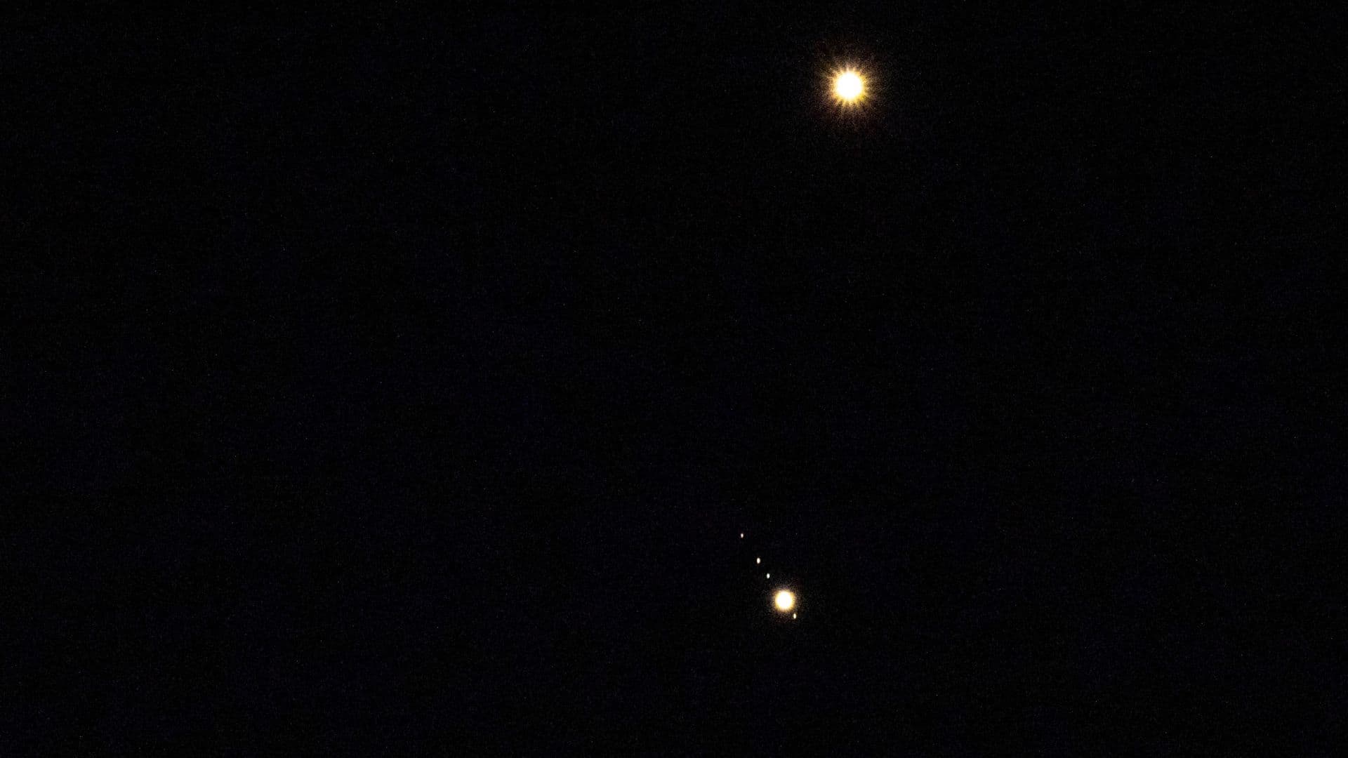 Venus is pictured shining brightly above Jupiter in a black sky. A number of small star-like objects can be seen in a line surrounding Jupiter, representing its four largest Galilean Moons.