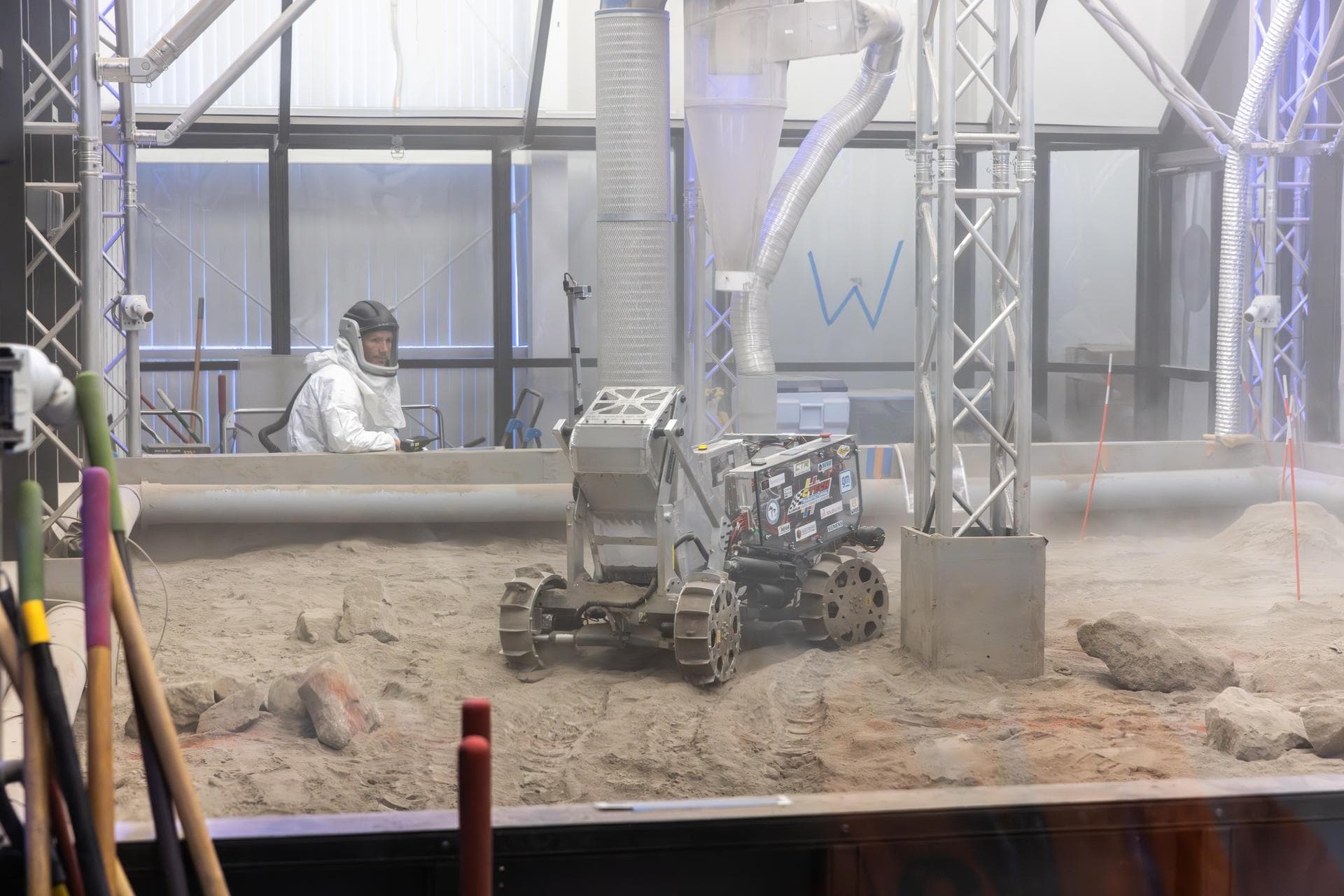 A person in a white cleanroom suit watches an excavating robot in a regolith-filled pit