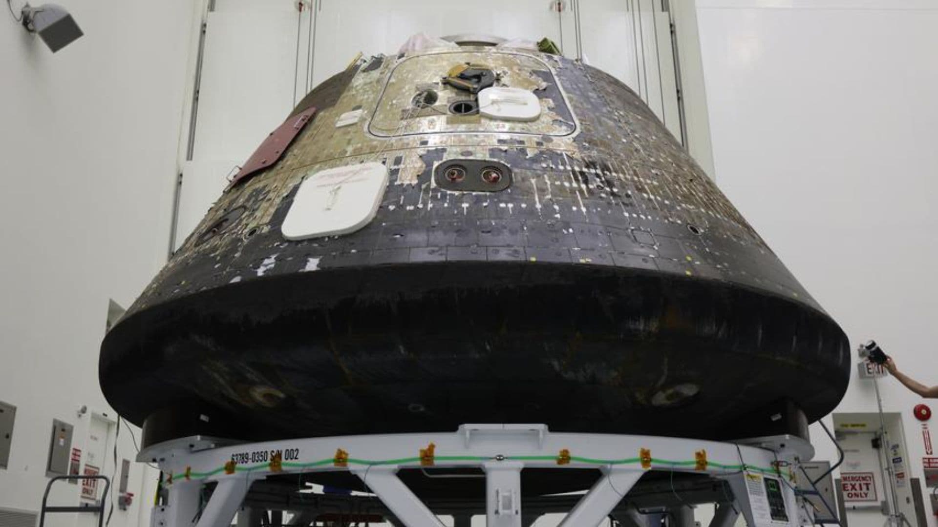 A space capsule with charred black on its bottom sits inside of a tall clean room.