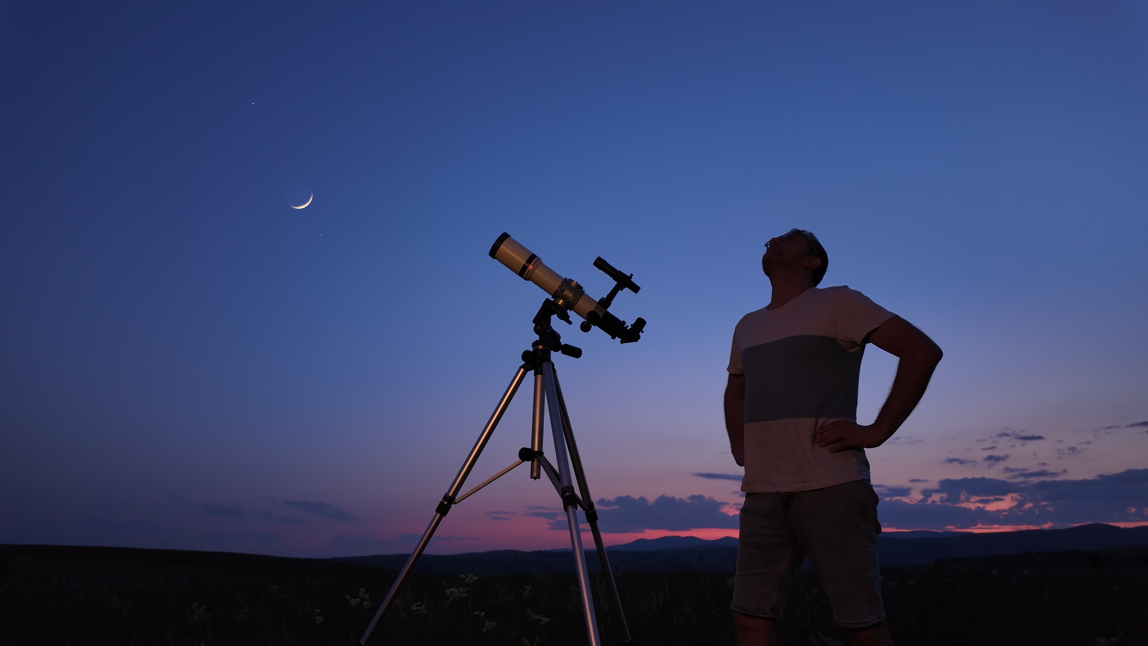 a man stands looking up the sky with his hands on his hips. there is a telescope to the left, looking up at a crescent moon.