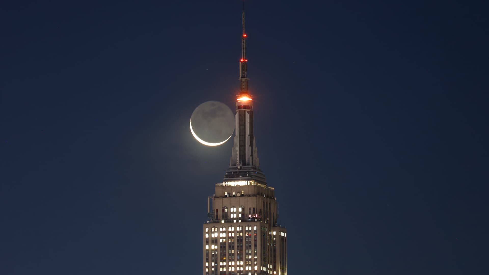 A thin crescent moon is shown next to the upper spire of a skyscraper in a dark sky. Its unlit surface is suffused with a soft glow, revealing the presence of lunar seas, or mare, on its surface.