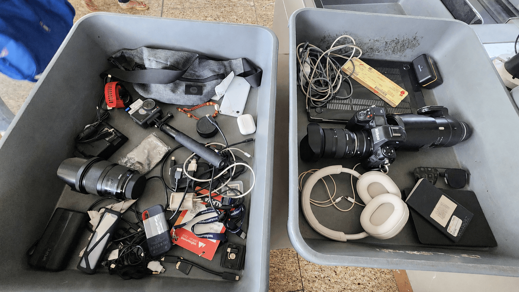 Two airport-security trays overflowing with a Nikon DSLR, an action cam, headphones, a sports watch, SSDs, batteries, and a tangle of cables
