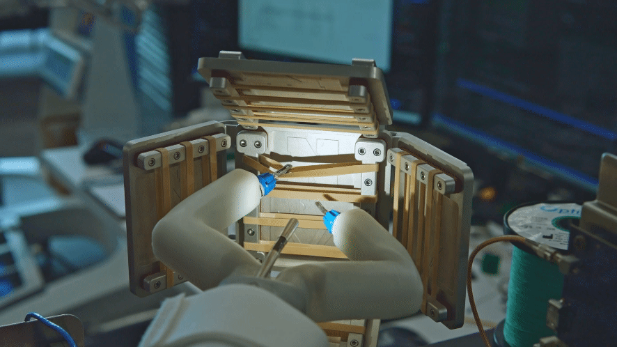 Two small white robotic arms grip a rubber band and snip it near the middle to simulate a surgical task. The robotic surgeon illuminates metallic panels with a small white light and uses one arm to stabilize the rubber band while the other cuts. There are many bands stretched and anchored inside of the compartment, while laboratory equipment and cables are softly blurred in the background.