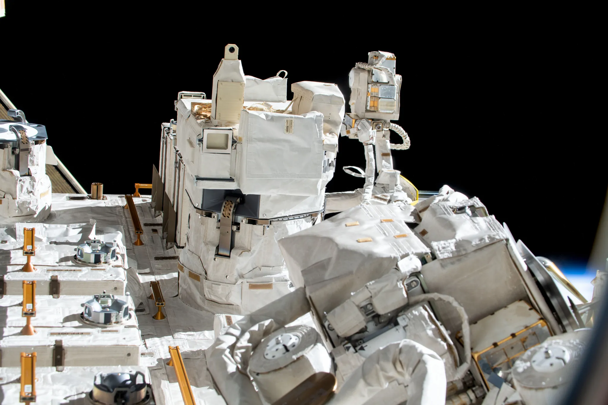 Box-shaped experiment hardware is covered by white thermal protection systems and mounted on the exterior of the International Space Station’s Japanese Experiment Module exposed facility. The exposed facility has many instruments, cables, and handrails along its structure. Near the top center of the image, the All Solid-state Lithium Ion Battery investigation appears as a rectangular unit that protrudes outward from the surrounding equipment.