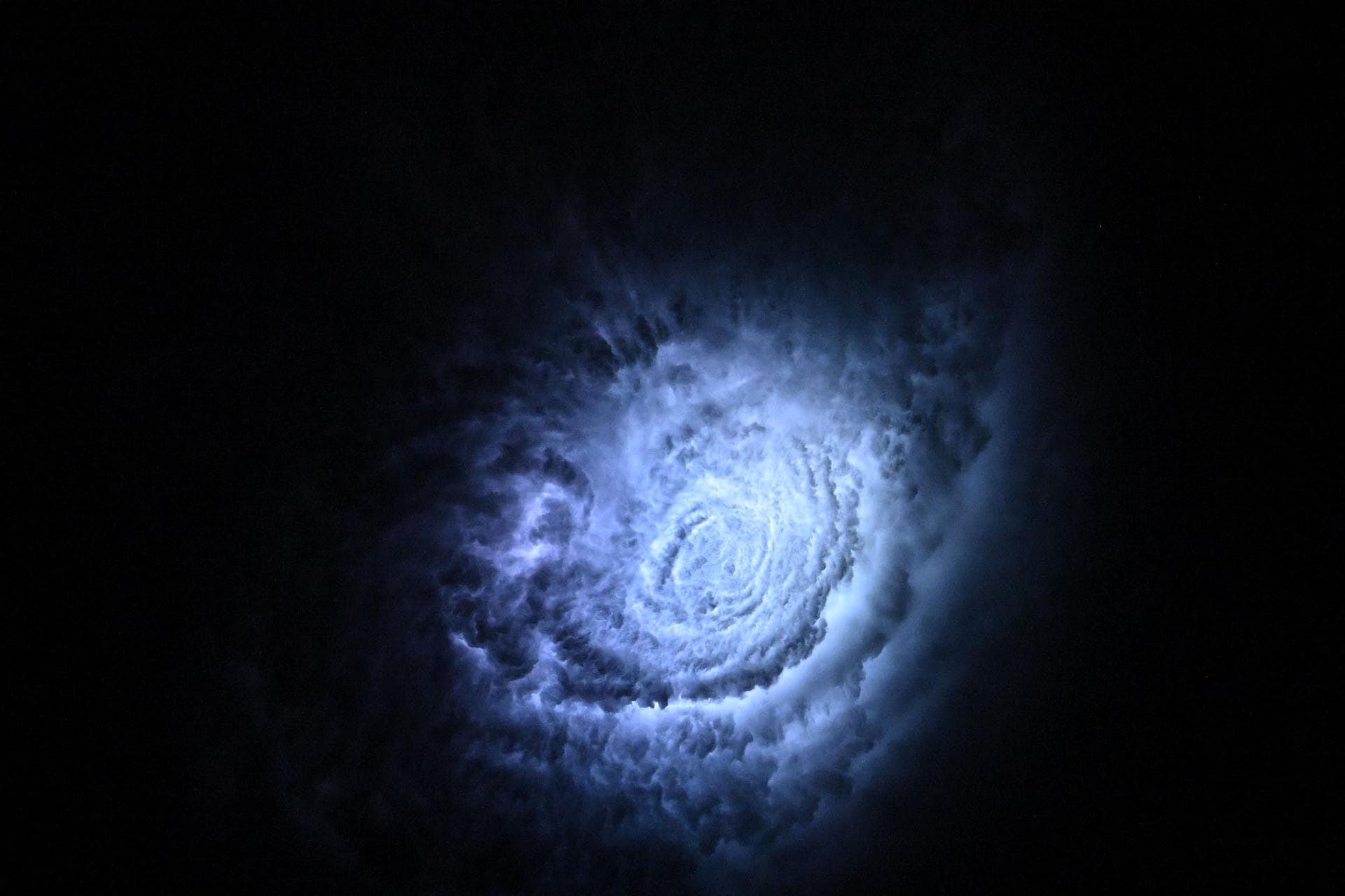 Blue lightning glows from within a large, spiraling mass of storm clouds over the Pacific coast of central Mexico, seen from space at night. The lightning flashes illuminate the cloud tops in pale blue-white tones, revealing a circular, ring-like structure like a glowing whirlpool.