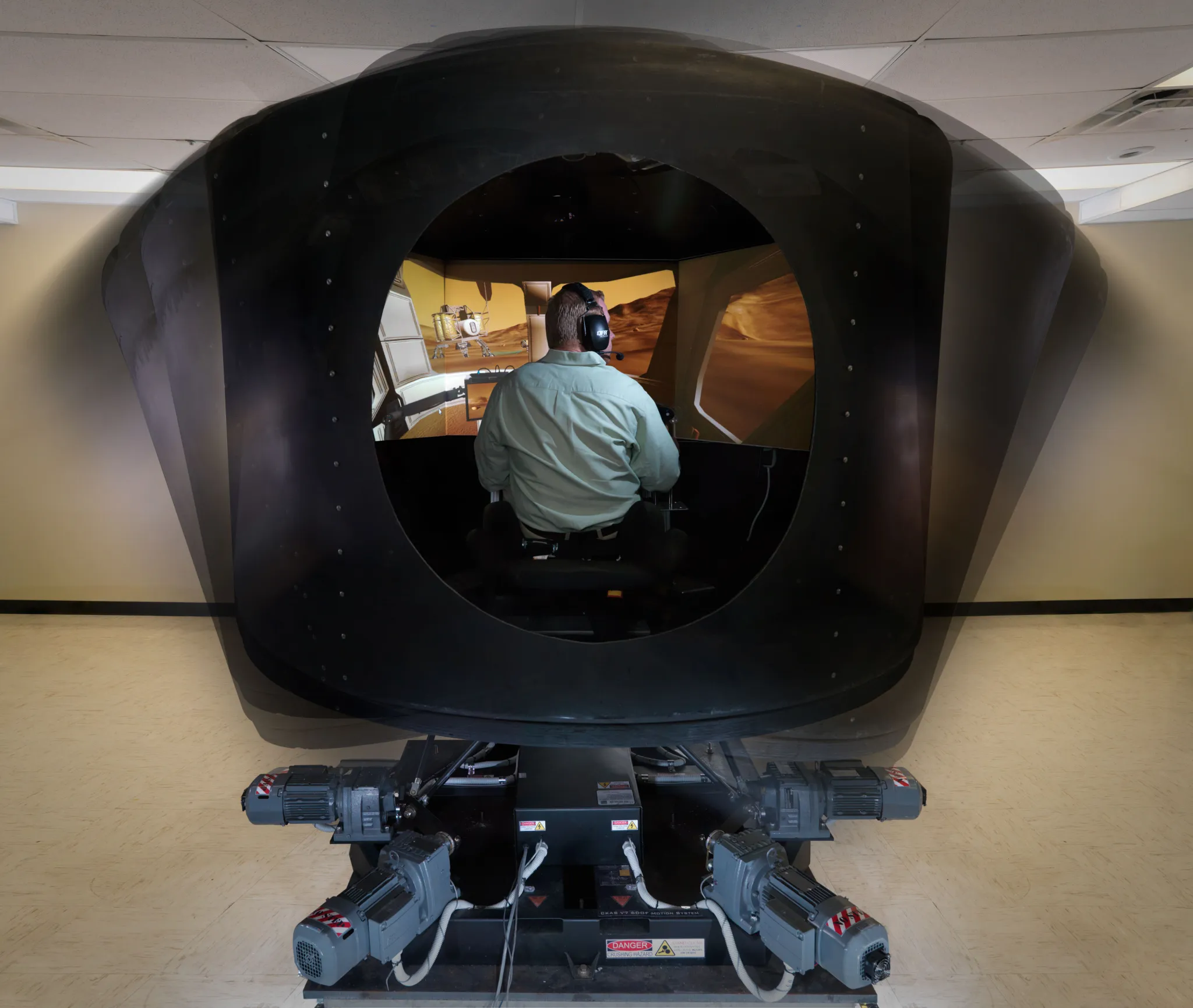 A test subject sits inside a large black motion simulator in a laboratory at NASA’s Johnson Space Center, wearing headphones and facing a curved screen displaying a spacecraft flight scene. The simulator surrounds the seated operator like a pod and is mounted on a mechanical base with visible motors and cables.