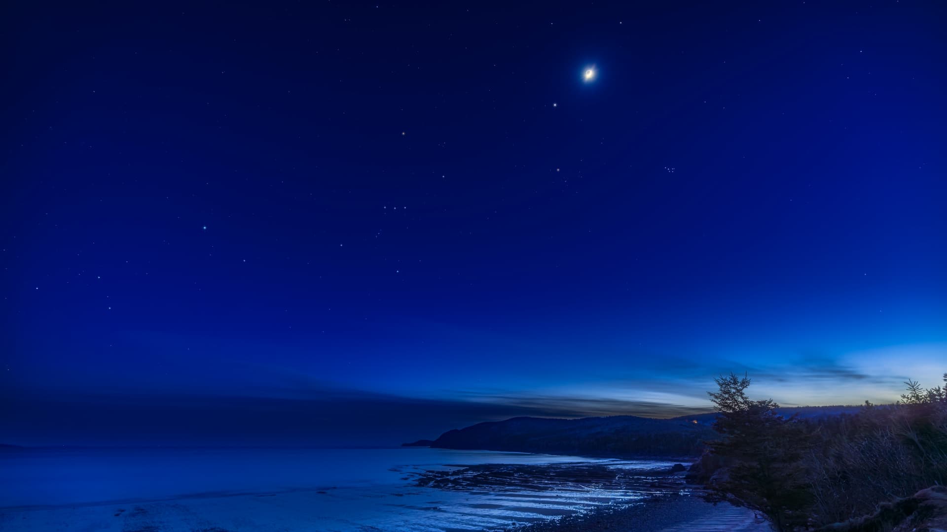 The moon is pictured glowing above the star-like form of Jupiter in a dark blue night sky above a snowy field bordering a road.