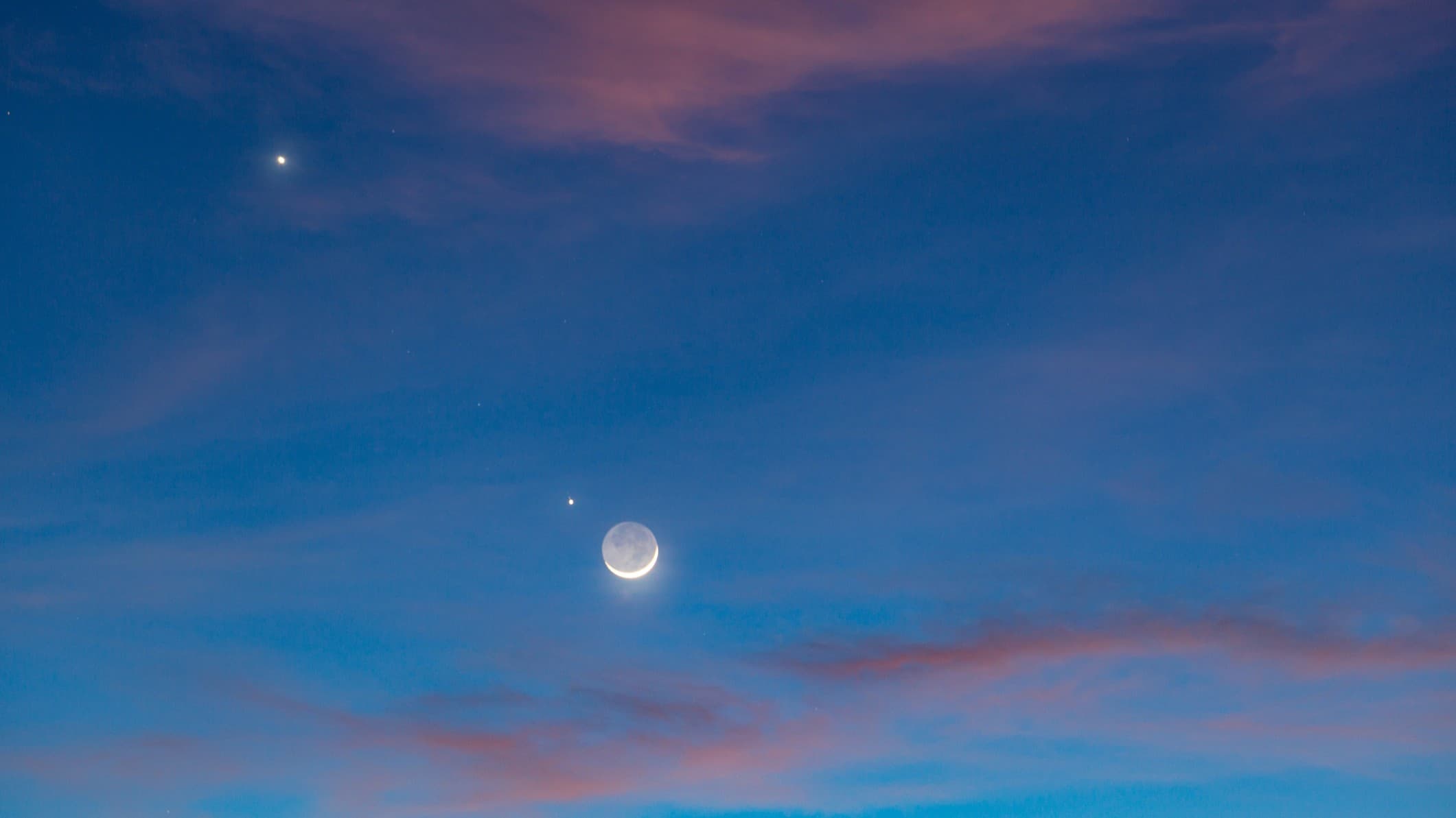 the thin crescent moon illuminated by earthshine sits in the dark blue sky alongside Venus and Jupiter