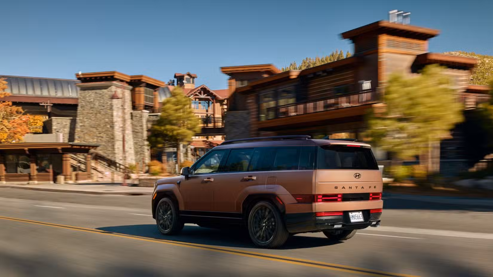 Dynamic rear 3/4 shot of a bronze 2025 Hyundai Santa Fe Hybrid driving through a city.