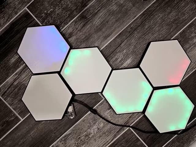 Hexagonal modular LED light panels connected together on a wooden floor, glowing in different colors with visible power and data cables.