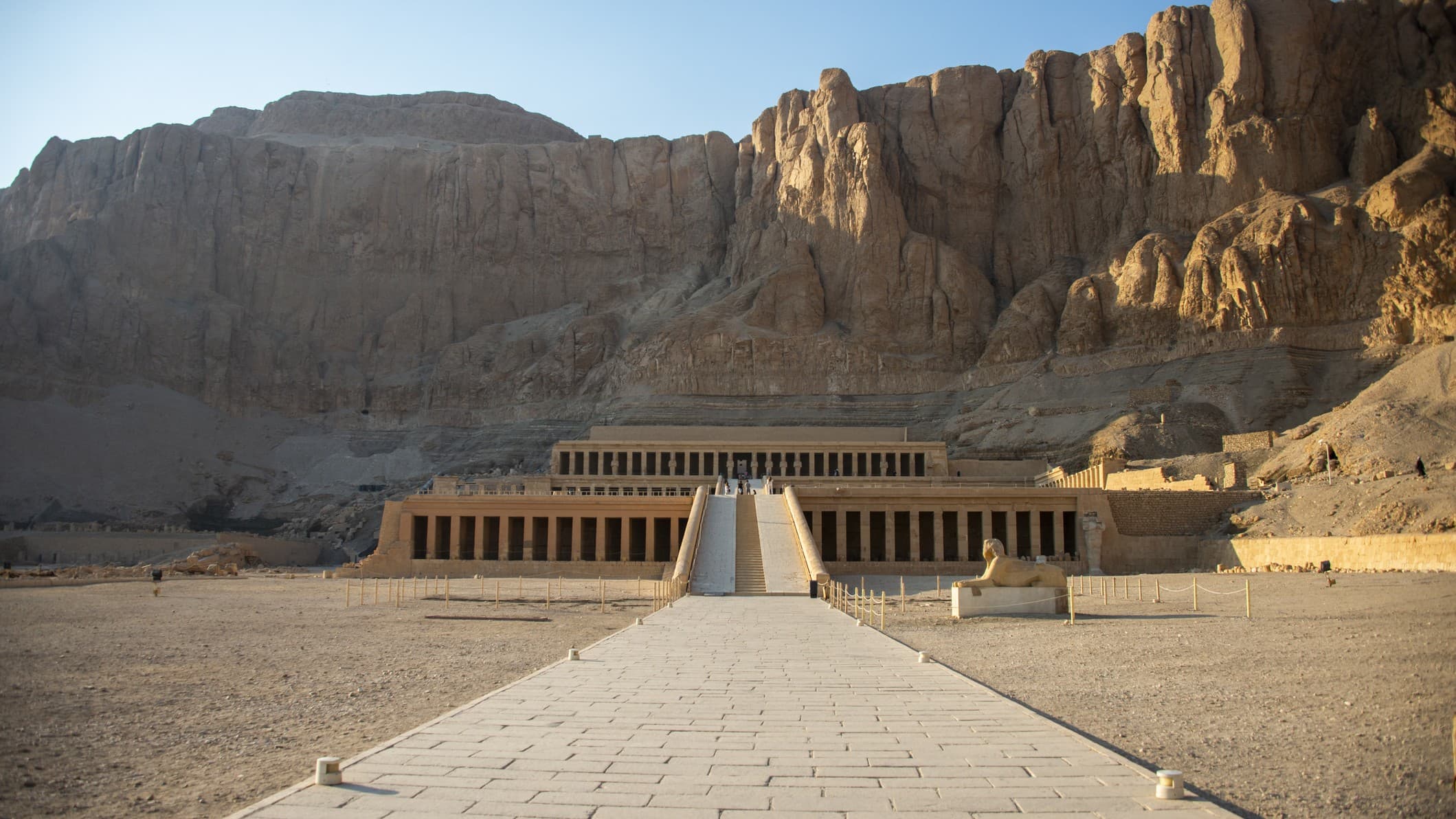 Queen Hatshepsut Temple is a large structure below towering rocks/cliff faces under an azure blue sky.