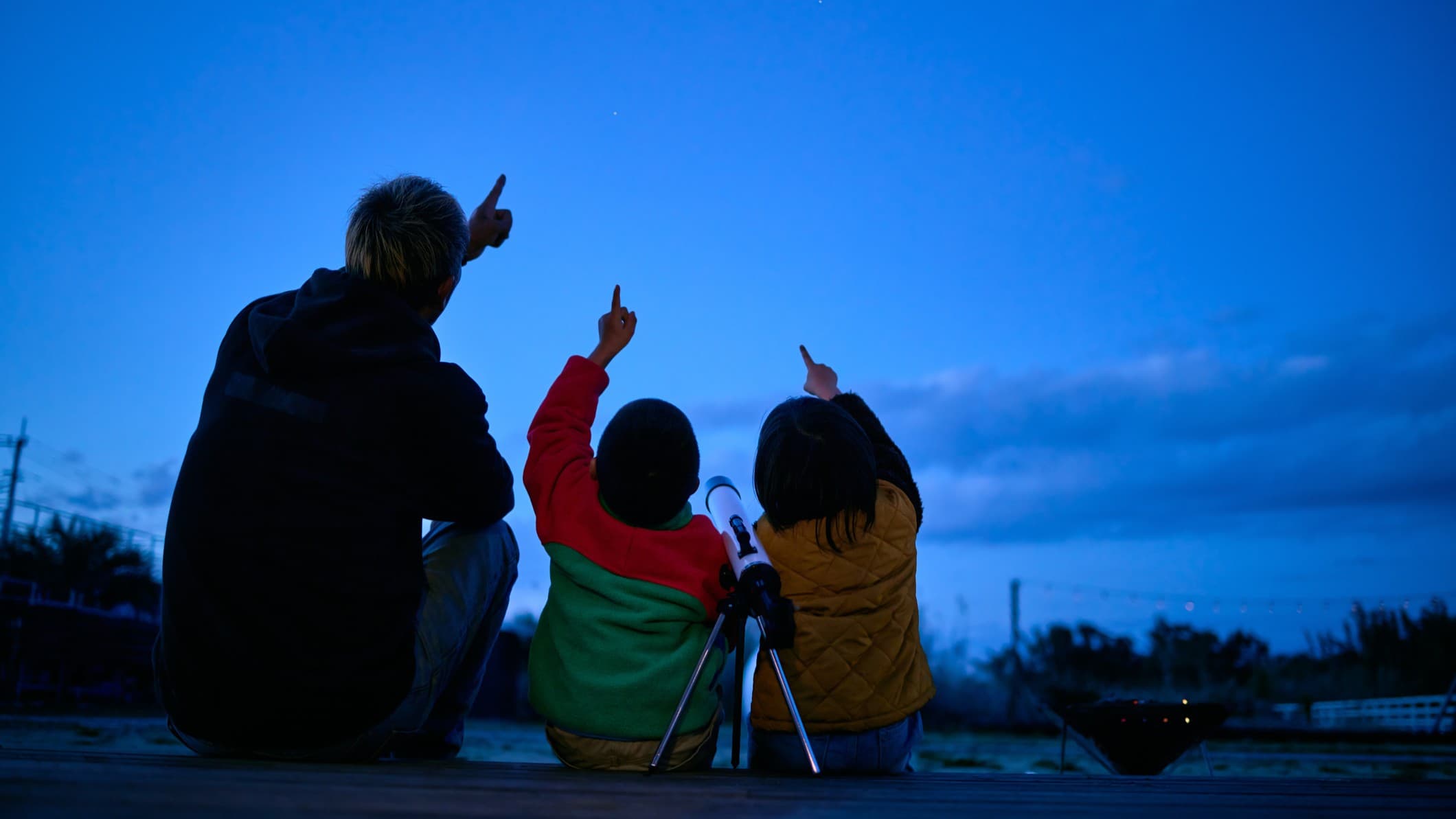 a man and two children sit and look at the night sky they are pointing up at the sky.