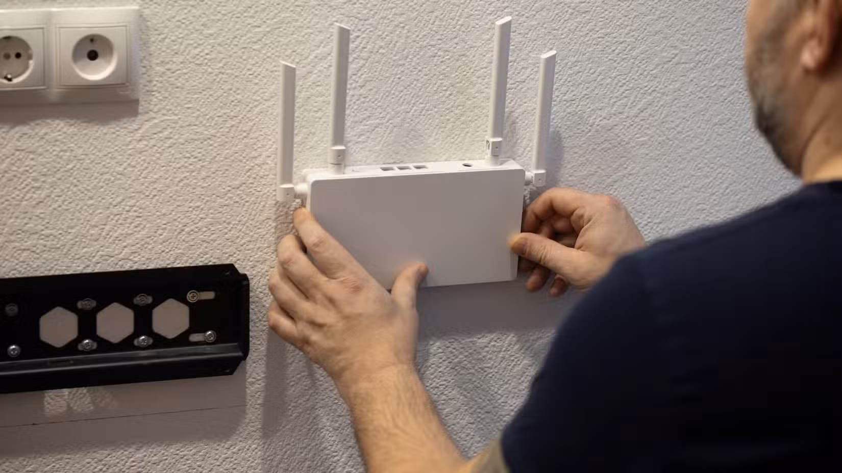 A man mounting a white Wi-Fi router on a white wall next to a TV mount.