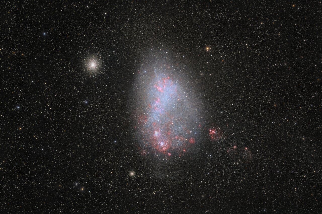 A black space background covered in stars has a central bright, egg-shaped galaxy with blue and red coloring.