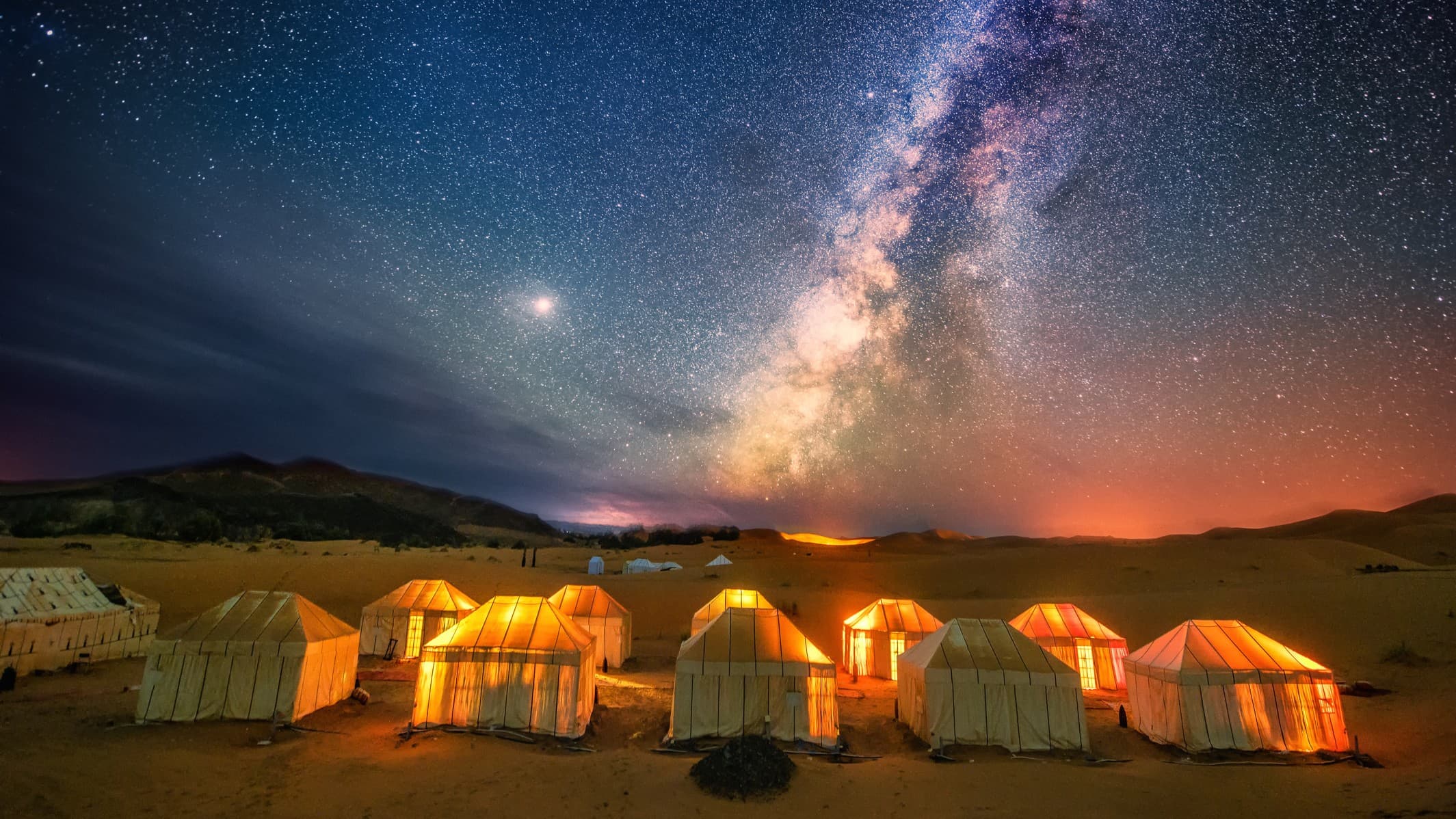 milky way galactic core stretching across the sky full of stars. there are tents in the foreground, bathed in soft golden glow from the lights within.
