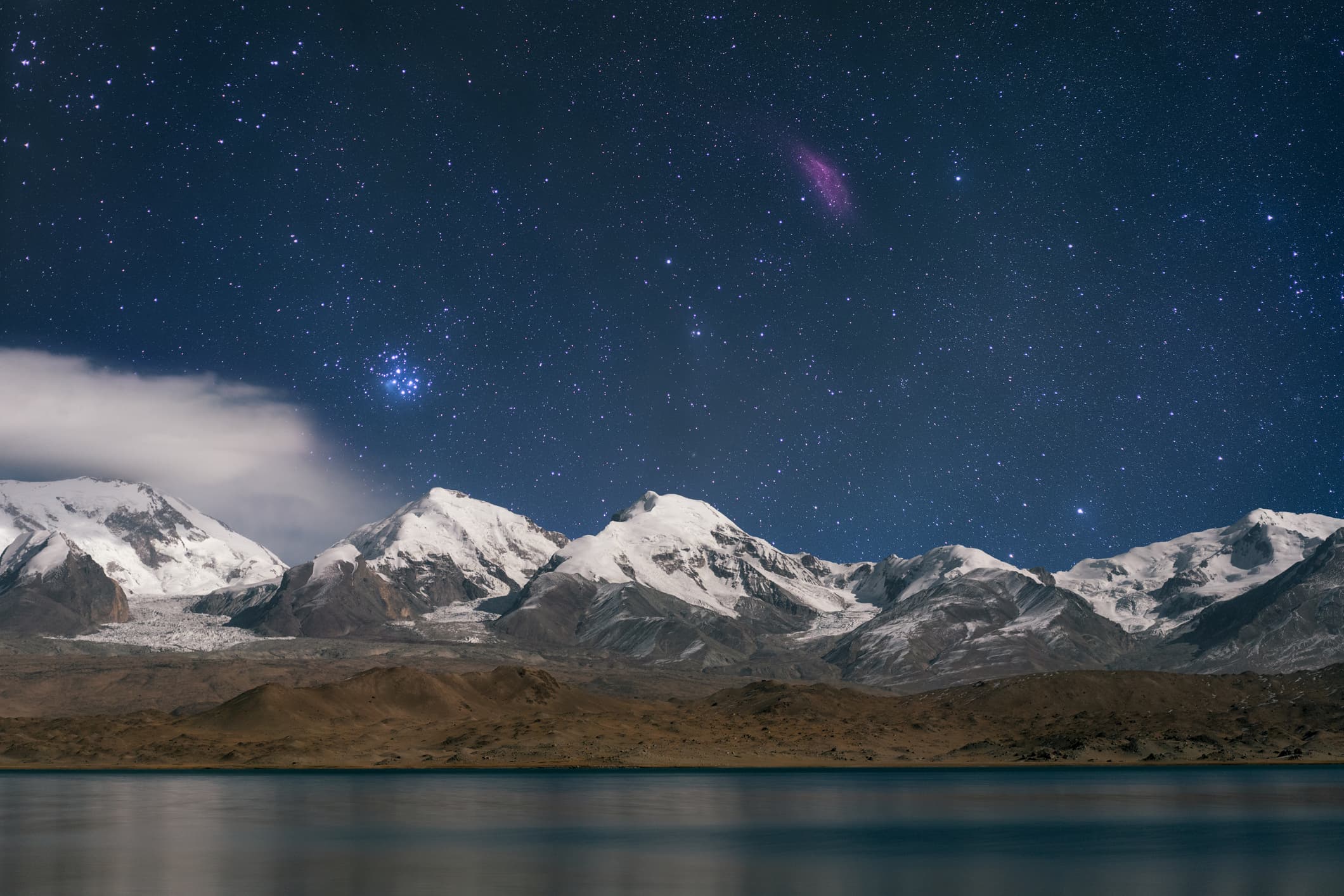 the Pleiades star cluster glistens as a patch of blue light above snow-capped mountains and water in the foreground below.