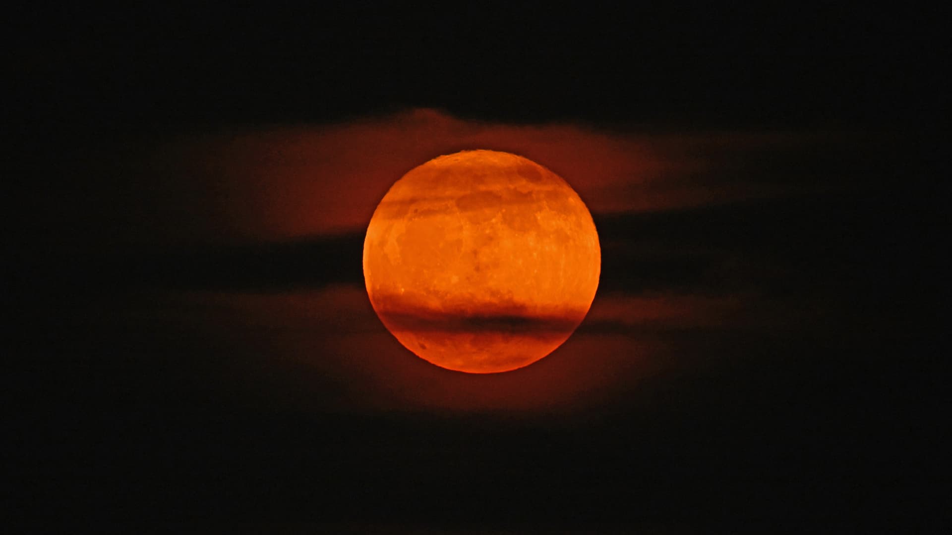 An orange-red moon glows in a dark night sky with streaks of cloud passing across its surface.