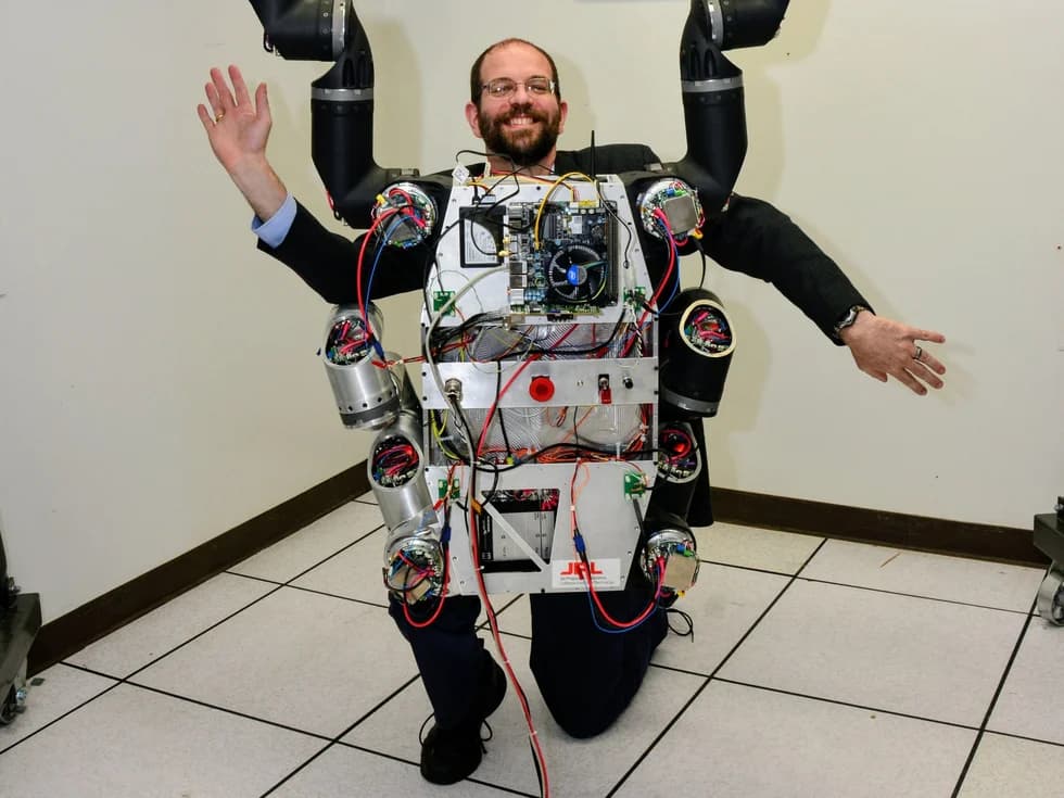 A smiling, bespectacled, bearded man kneels posed behind a robotic torso.