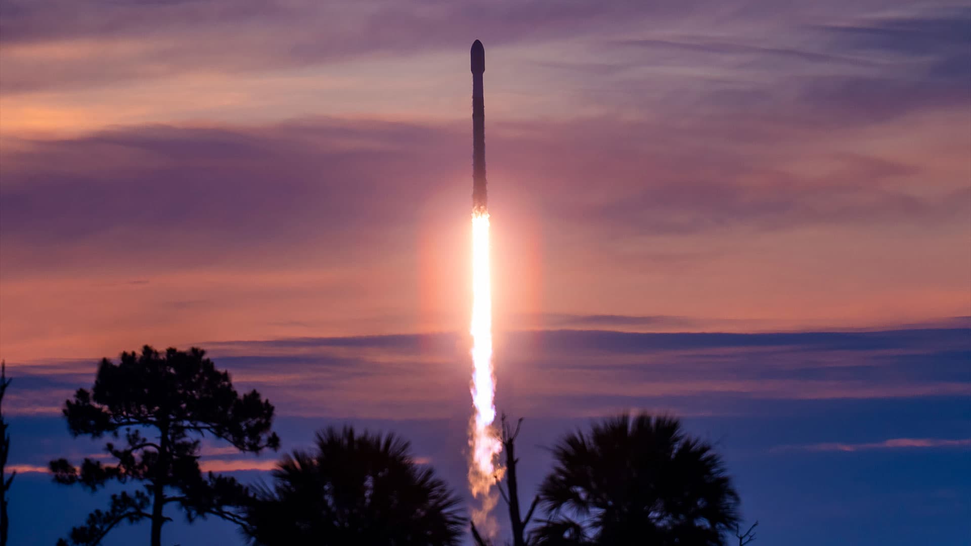 A rocket lifts off into the dawn sky, set against purple clouds and a pinkish-orange horizon