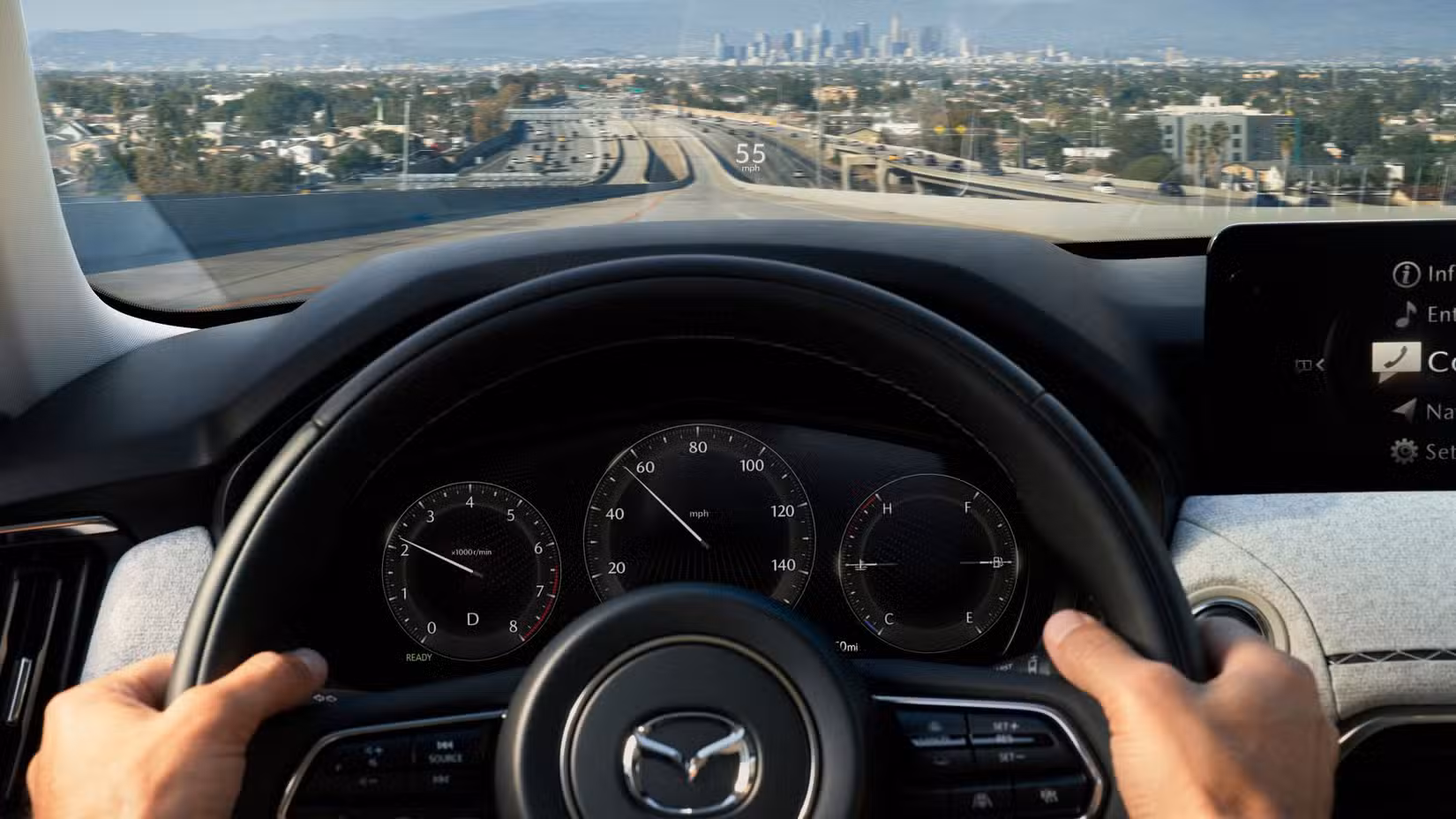 Close-up shot of a man's hands on the steering wheel of a 2025 Mazda CX-90, with a citscape in the background.