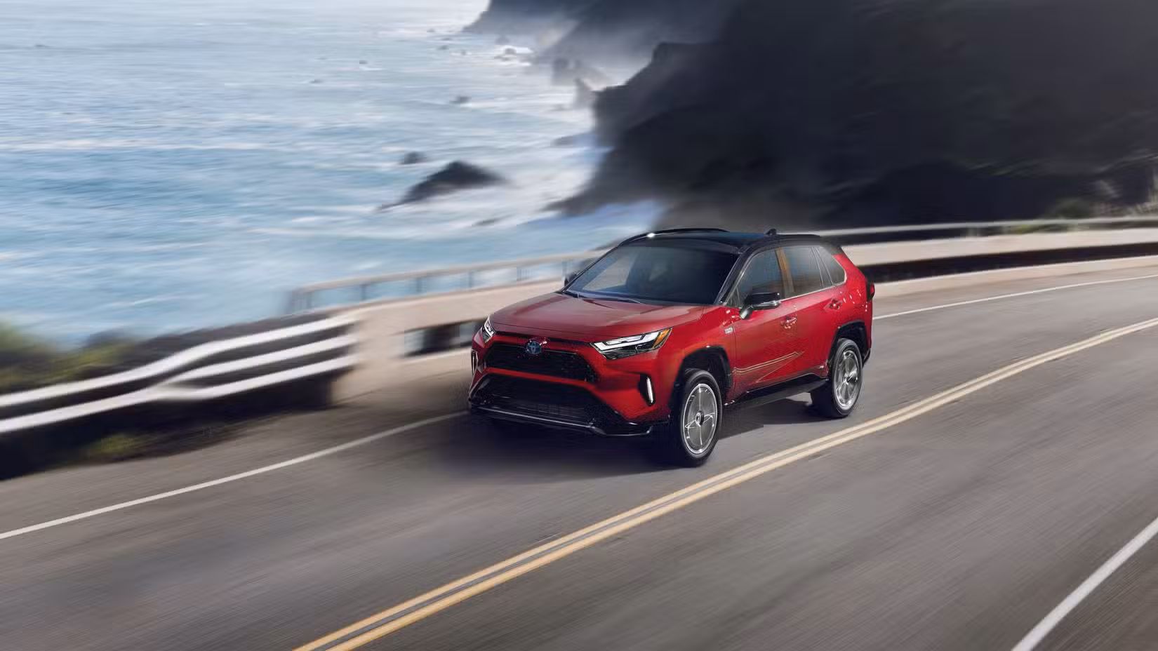 Dynamic front 3/4 shot of a red 2024 Toyota RAV4 Prime XSE driving on a road running beside the ocean.