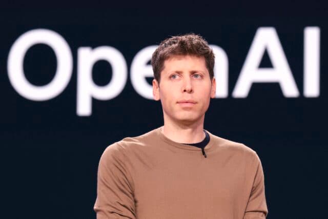 OpenAI CEO Sam Altman standing in front of a backup with a large OpenAI logo.